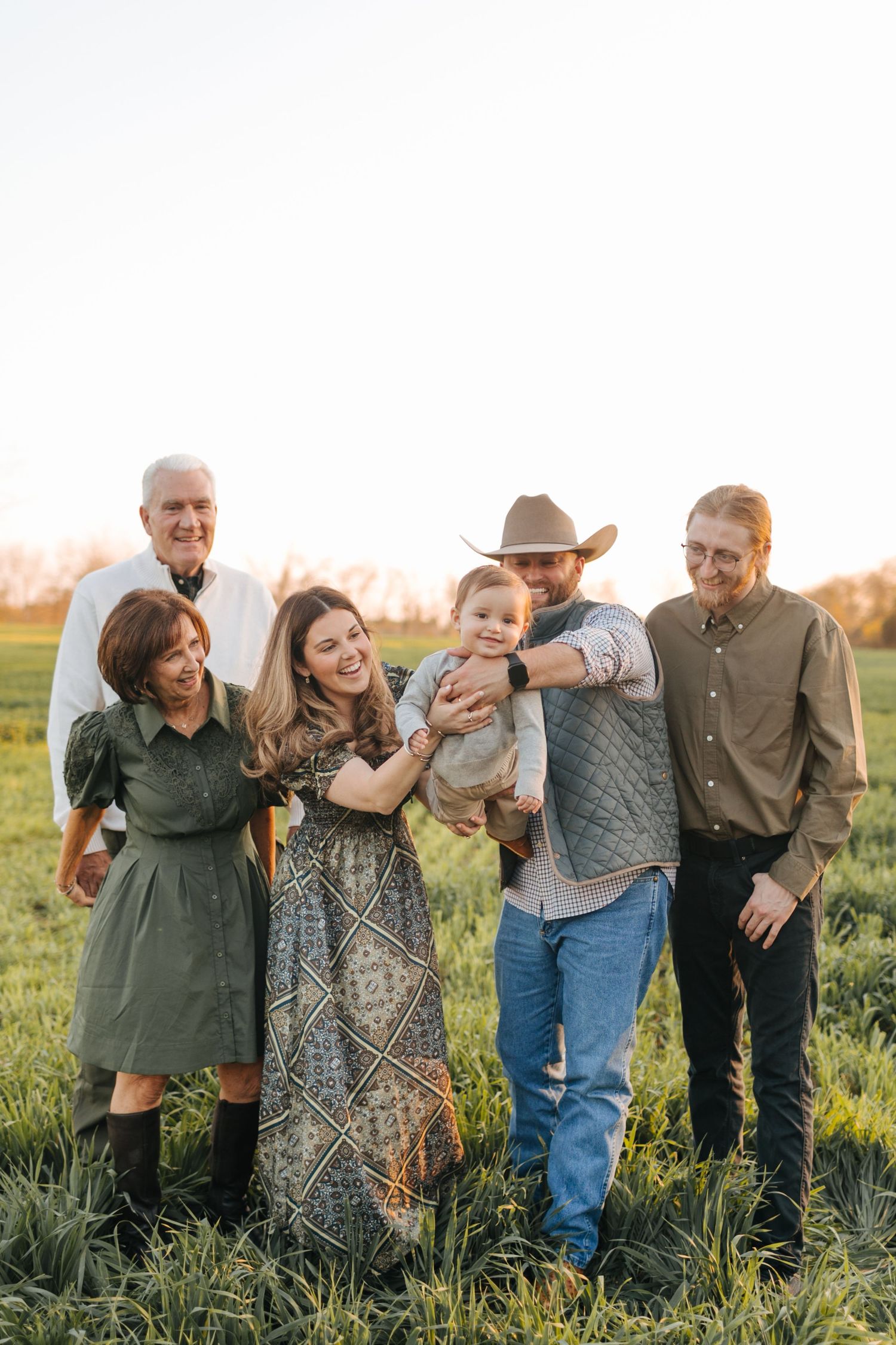 Extended family laughing and playing with baby in green field, bathed in warm golden hour sunlight.