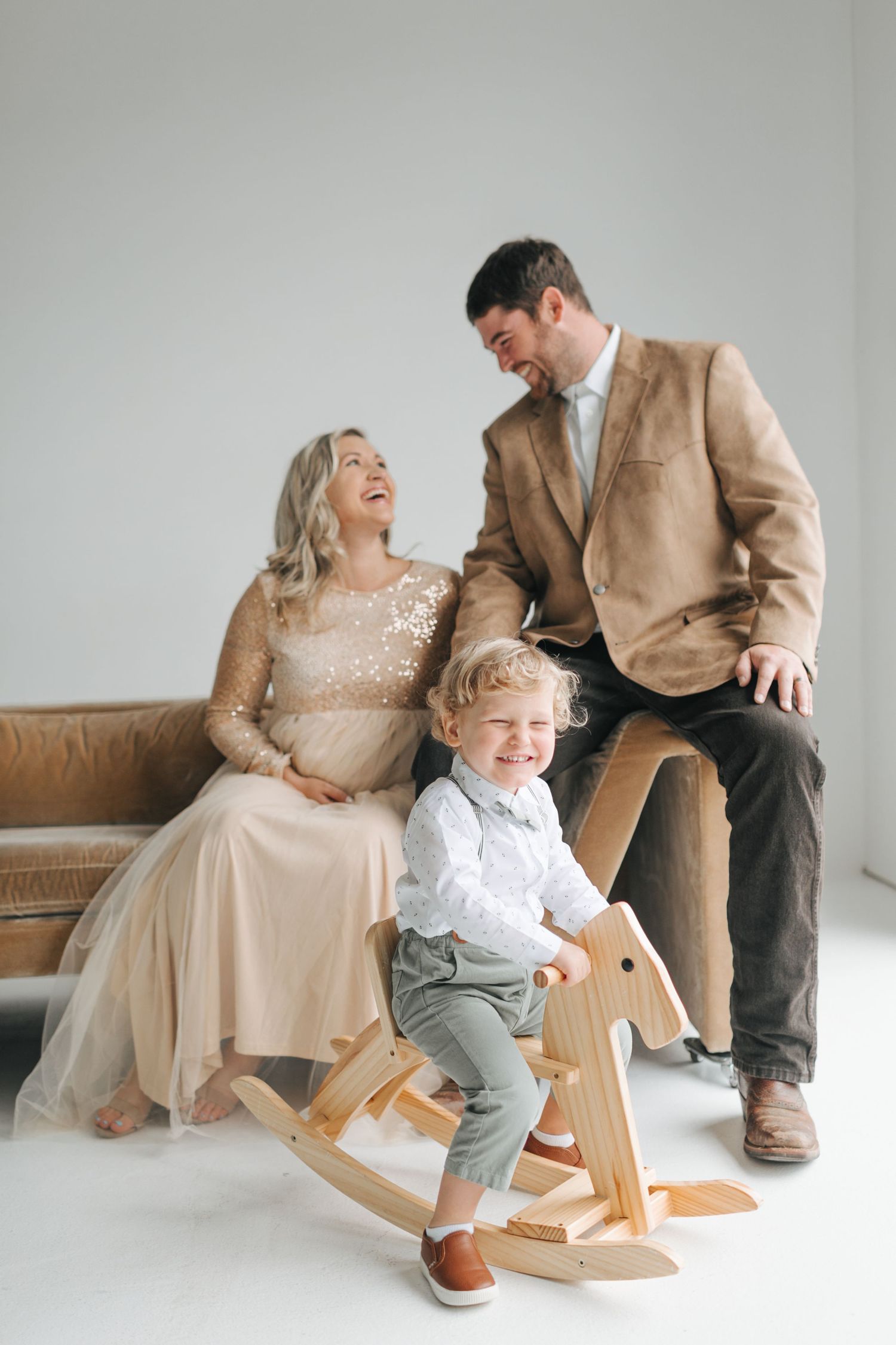 A happy family with a pregnant mother in beige dress, father in tan blazer, and toddler on a wooden rocking horse.