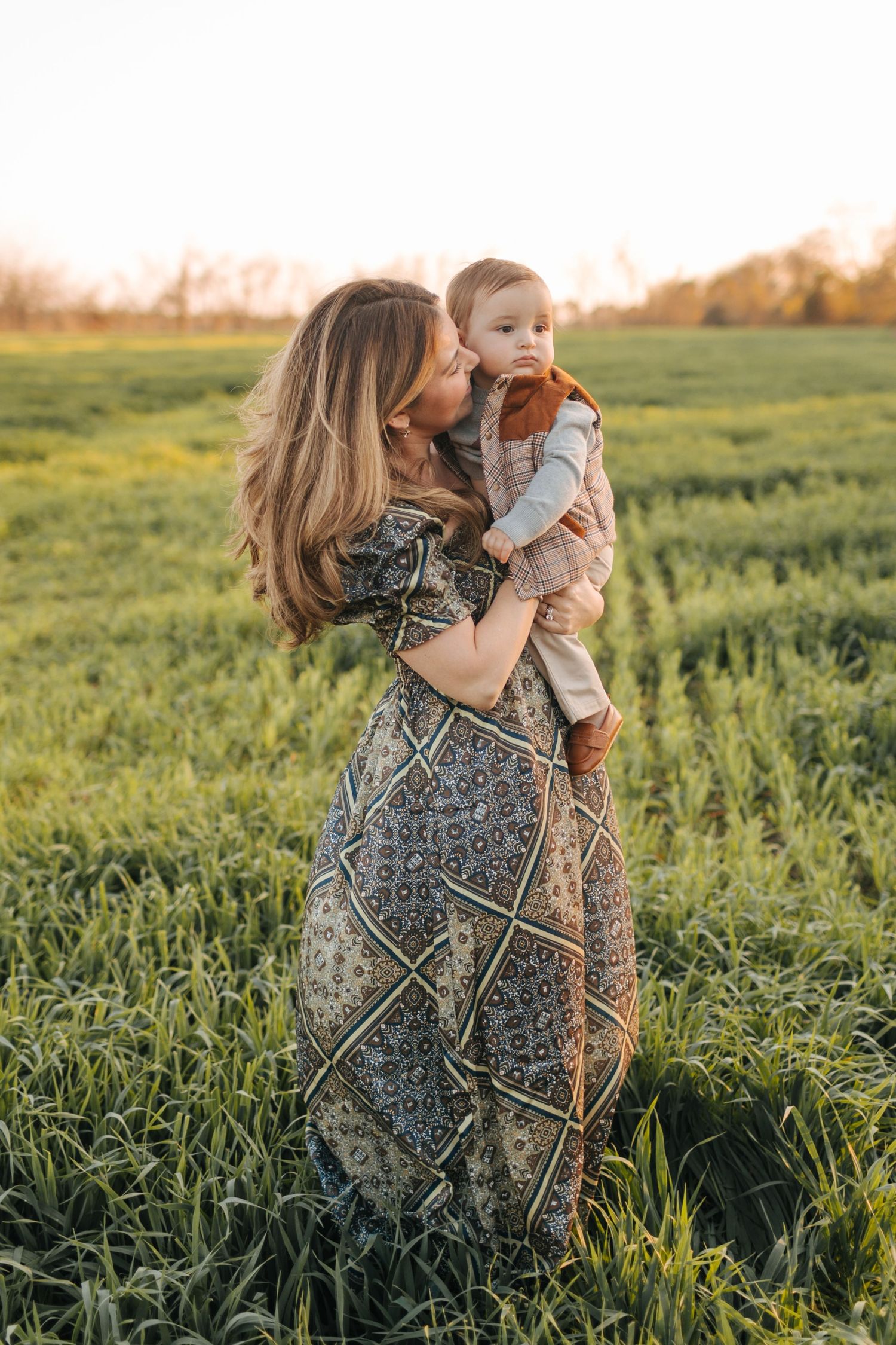 Young mother in boho maxi dress kissing her baby while standing in a lush green field at sunset.