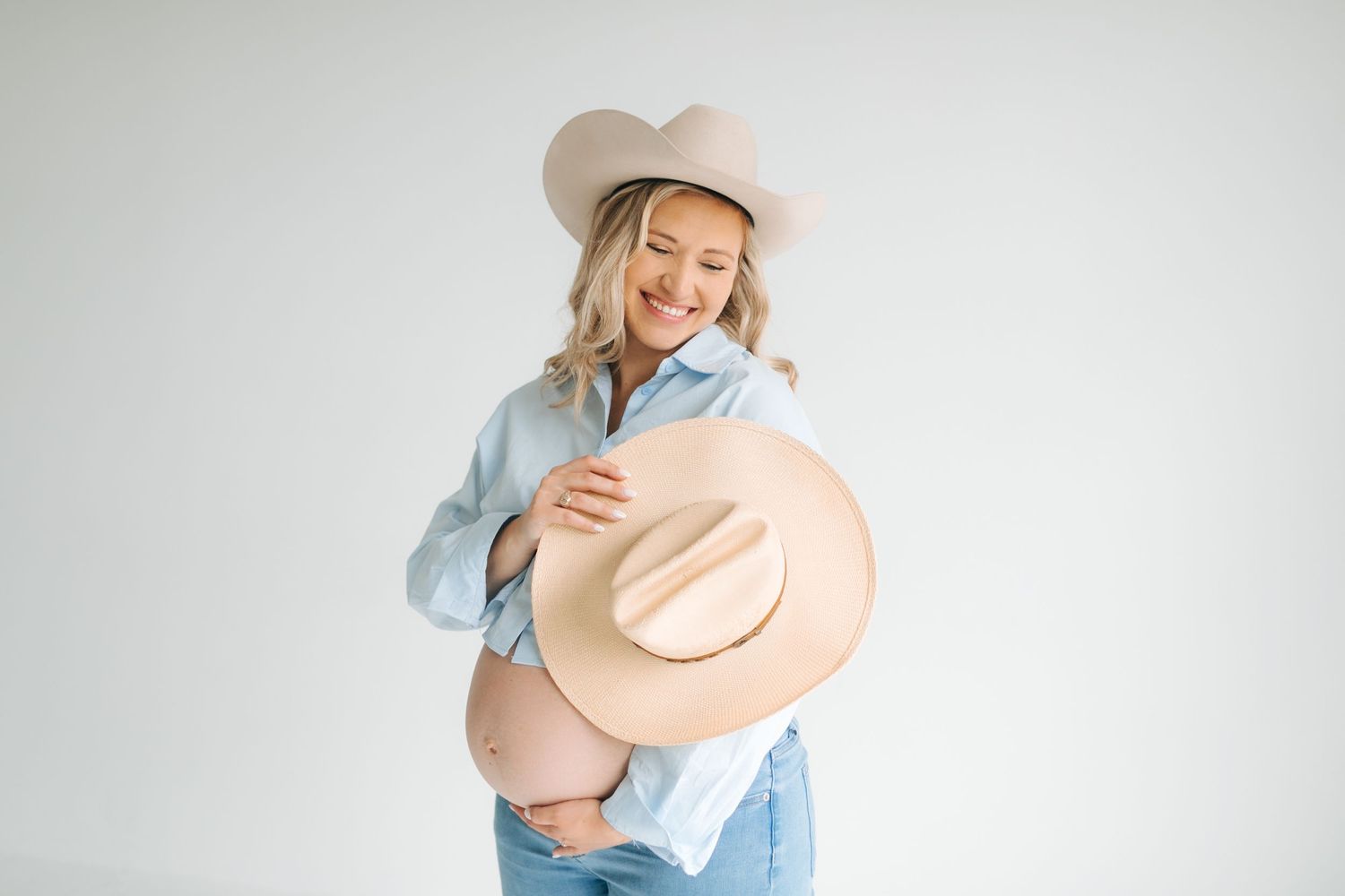 Pregnant woman in denim shirt and cowboy hat holding belly in studio portrait.