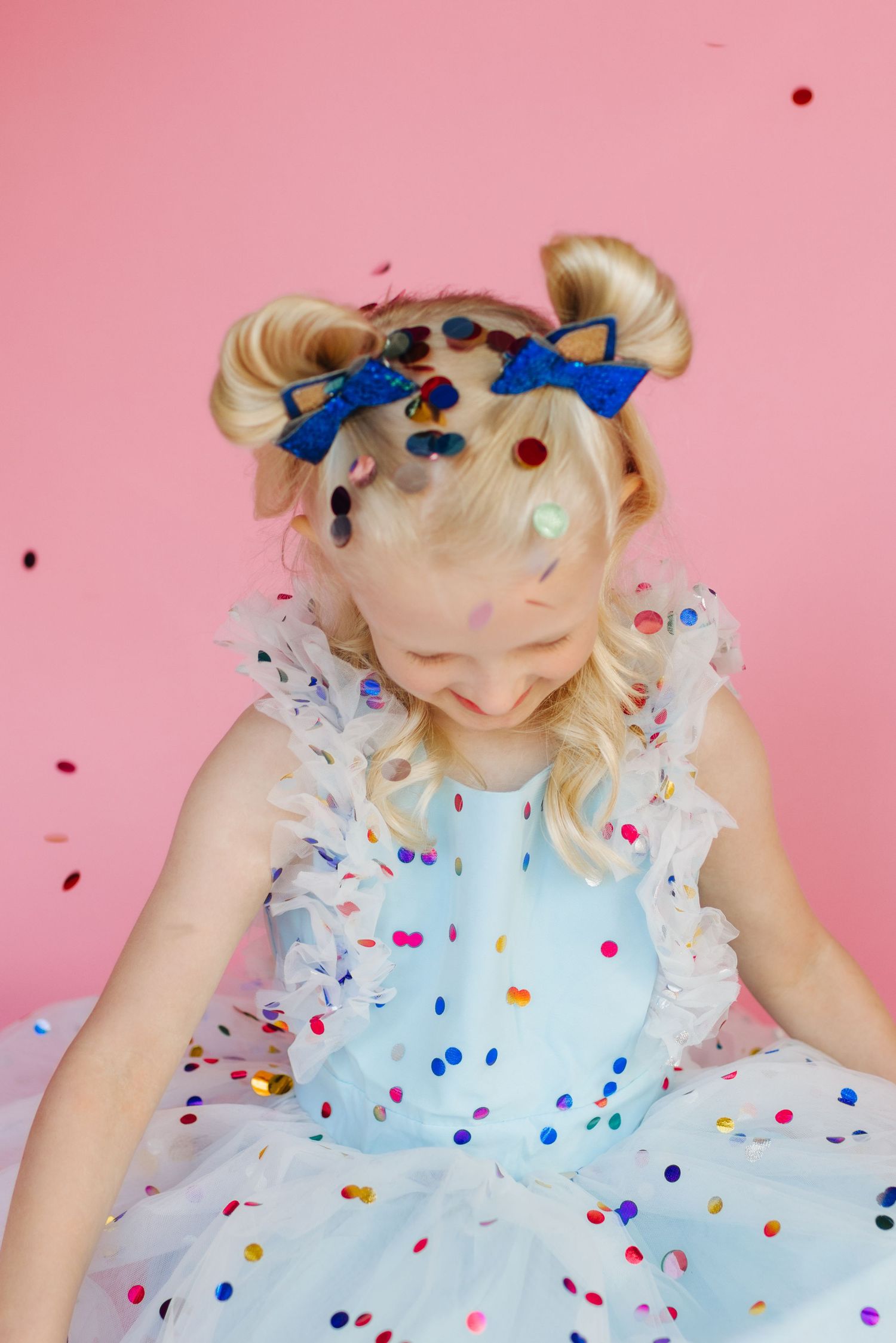 A joyful blonde girl in a light blue tutu dress with blue bows, surrounded by colorful falling confetti on a pink background.