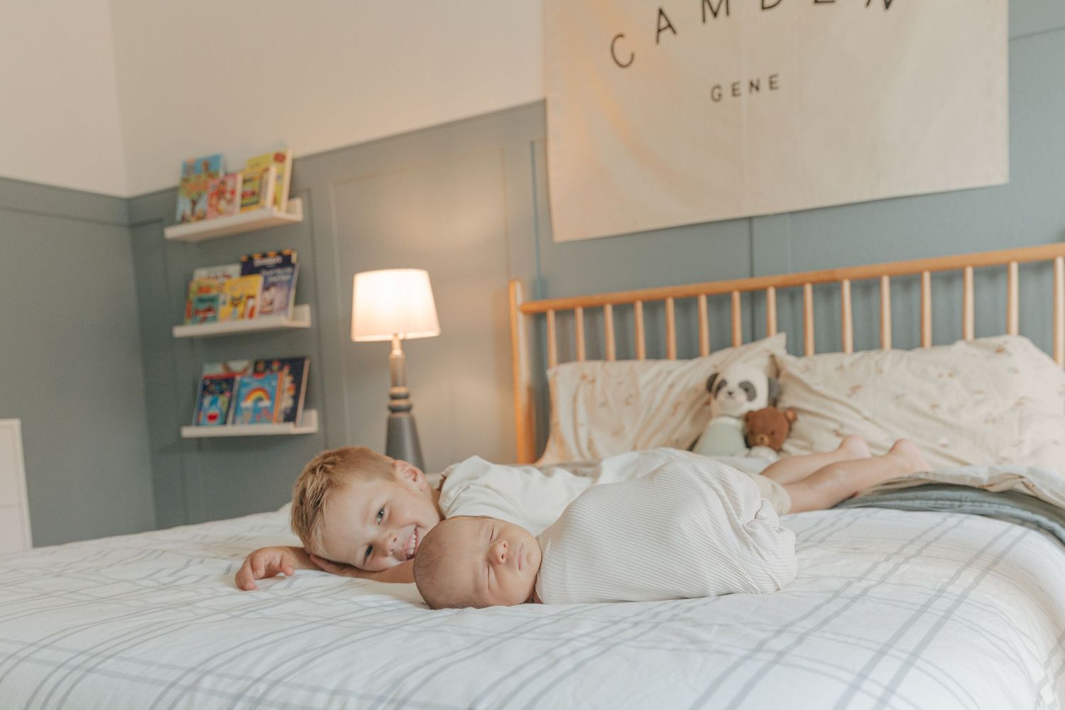Young child cuddles newborn baby on bed in cozy nursery with Camden Gene banner on wall.