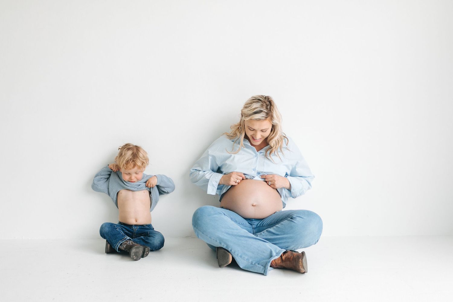 A pregnant woman in denim sits cross-legged with her toddler son, both lifting their shirts to show their bellies.