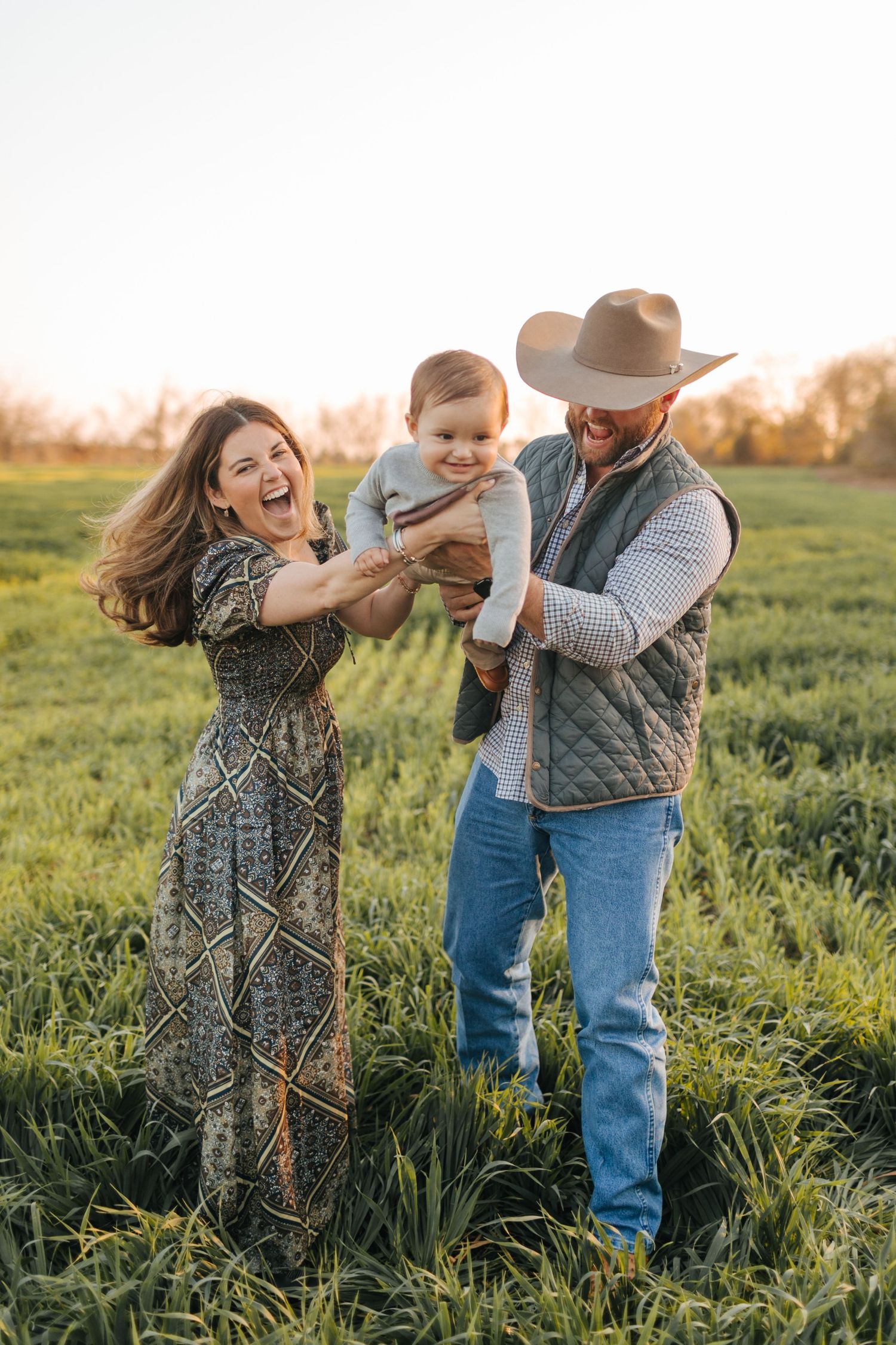 Laughing family with joyful baby in green field at sunset, mom in maxi dress, dad in cowboy hat holding child.