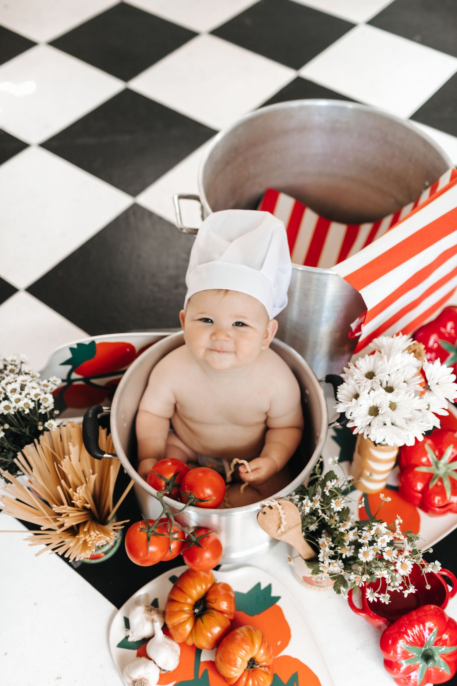 Adorable baby wearing chef hat sitting in large pot surrounded by tomatoes, flowers, and vegetables on checkered floor.