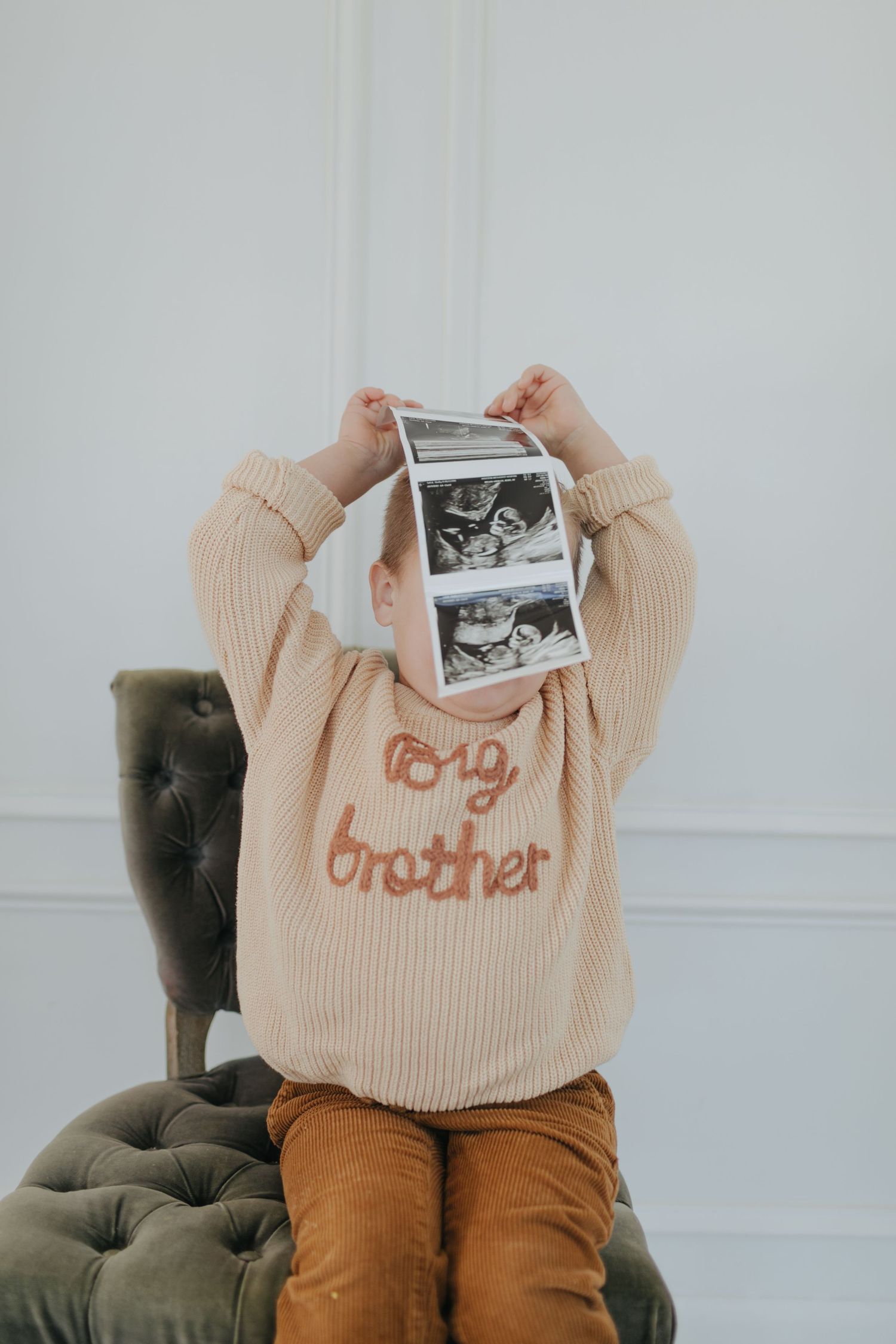 A toddler in a big brother sweater holds ultrasound photos above their face while sitting on a green velvet chair.