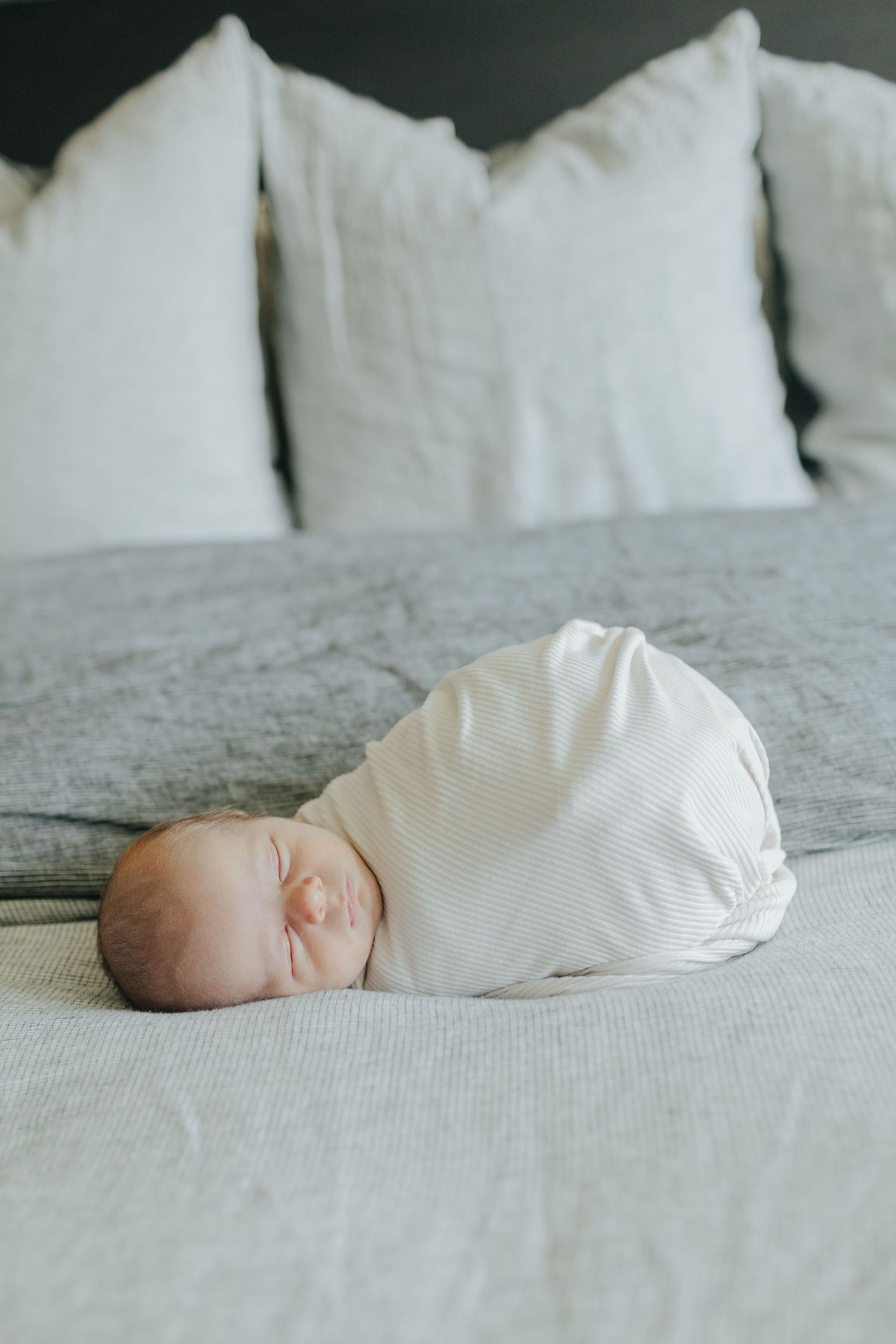 Peaceful newborn baby swaddled in white sleeping on a gray linen bed with white pillows.