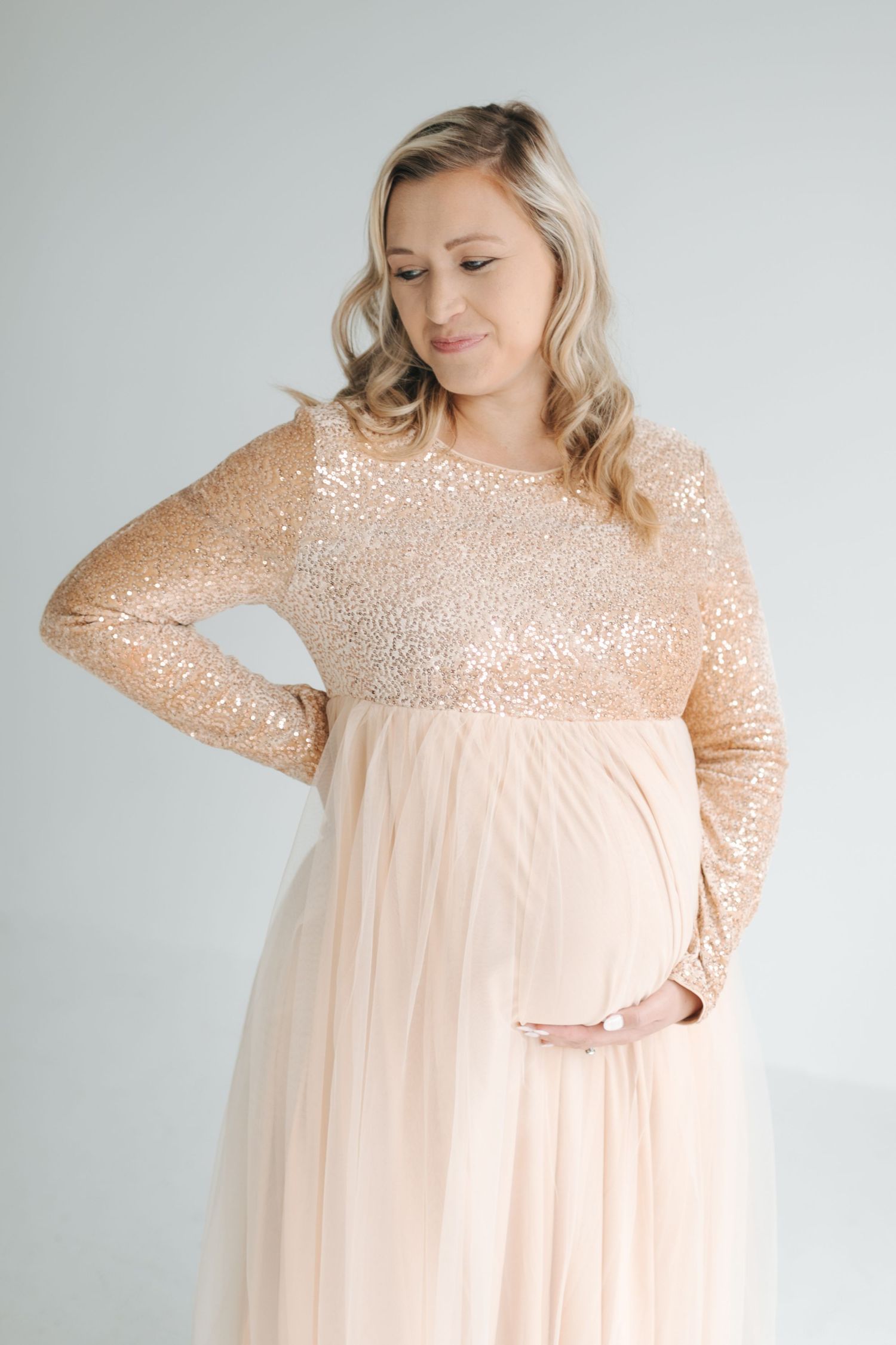 Pregnant woman in rose gold sequin maternity gown cradling baby bump in studio.