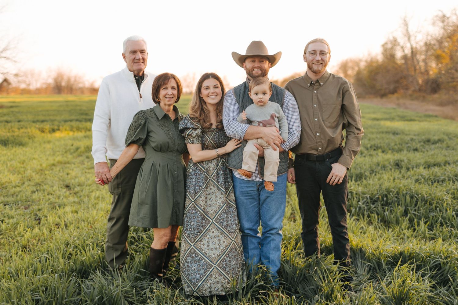 Extended family of six smiling together in green farm field at golden hour in country-style attire.