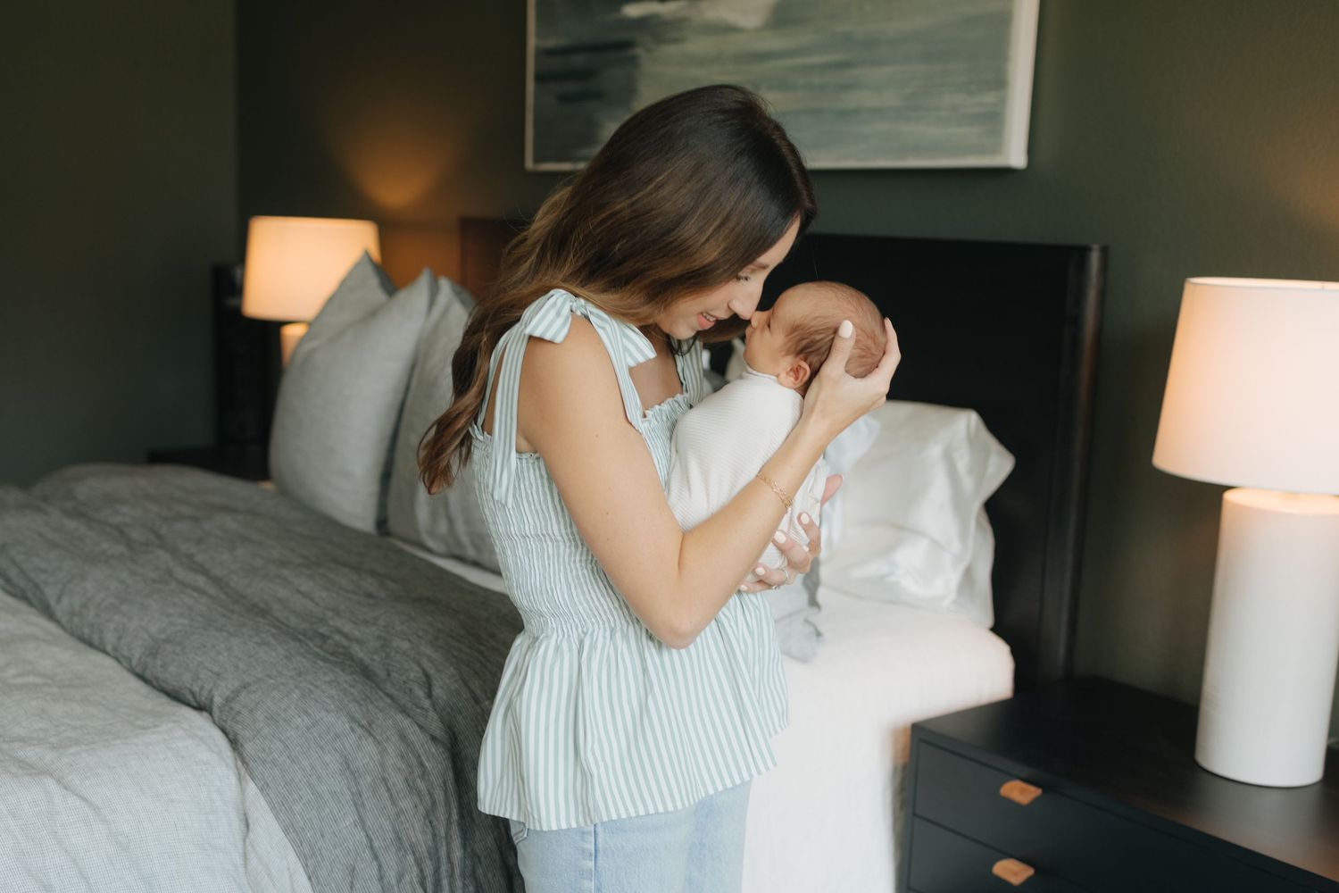 A mother tenderly holds her newborn baby in a softly lit bedroom with gray bedding and warm lamps.