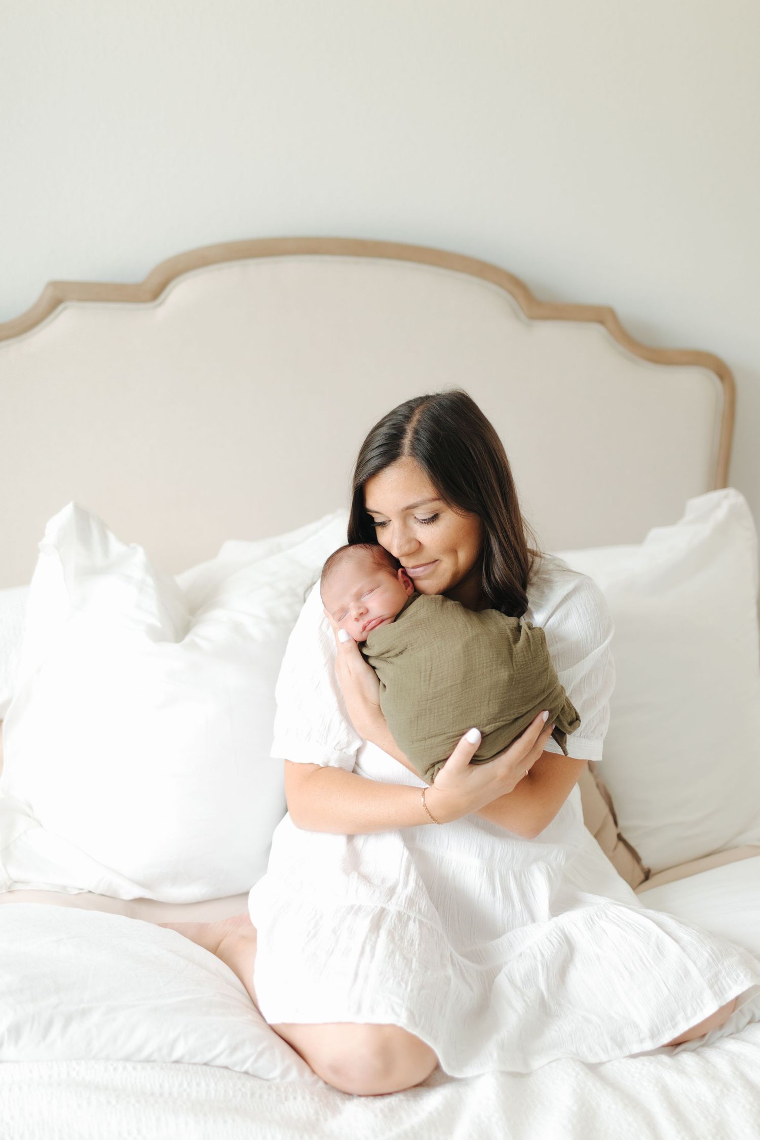 Dark-haired mother in white dress on white bed peacefully cradling sleeping olive-wrapped newborn baby.