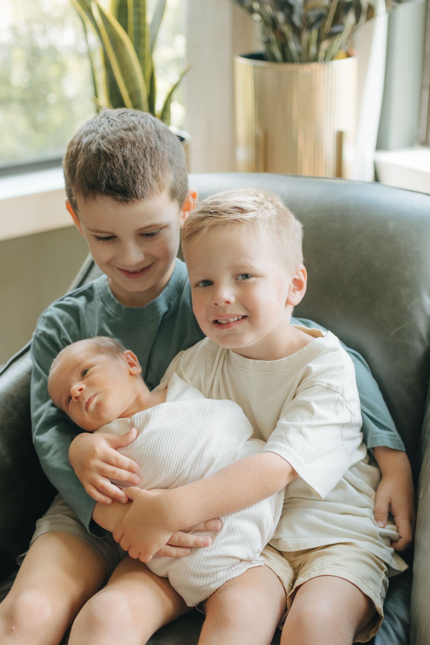 Two young brothers sitting in a chair, lovingly holding their sleeping newborn baby sibling.