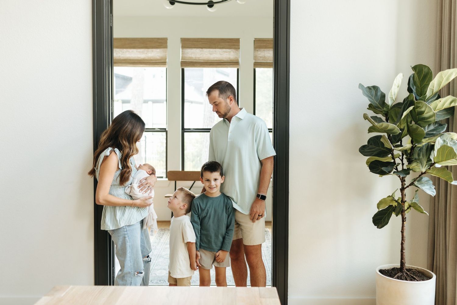 Young family with two children standing together in a bright modern home entryway with a fiddle leaf fig plant.