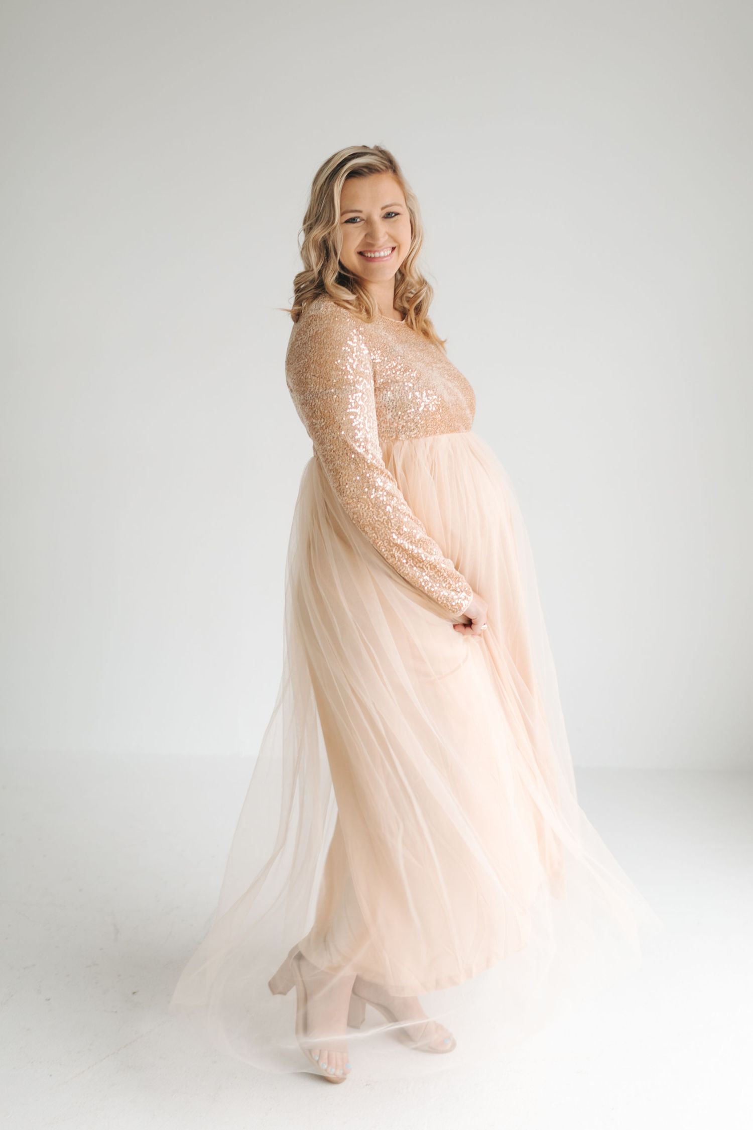 Pregnant woman in sequin and tulle blush gown smiling in bright white studio.