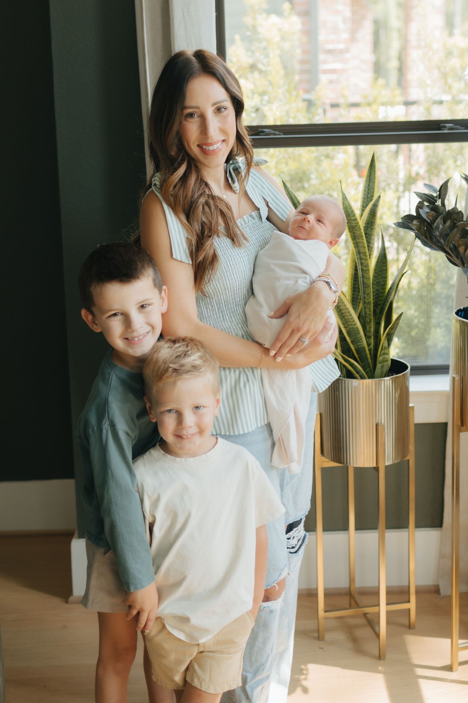 Smiling mother holding newborn baby while posing with two young sons near a large window.