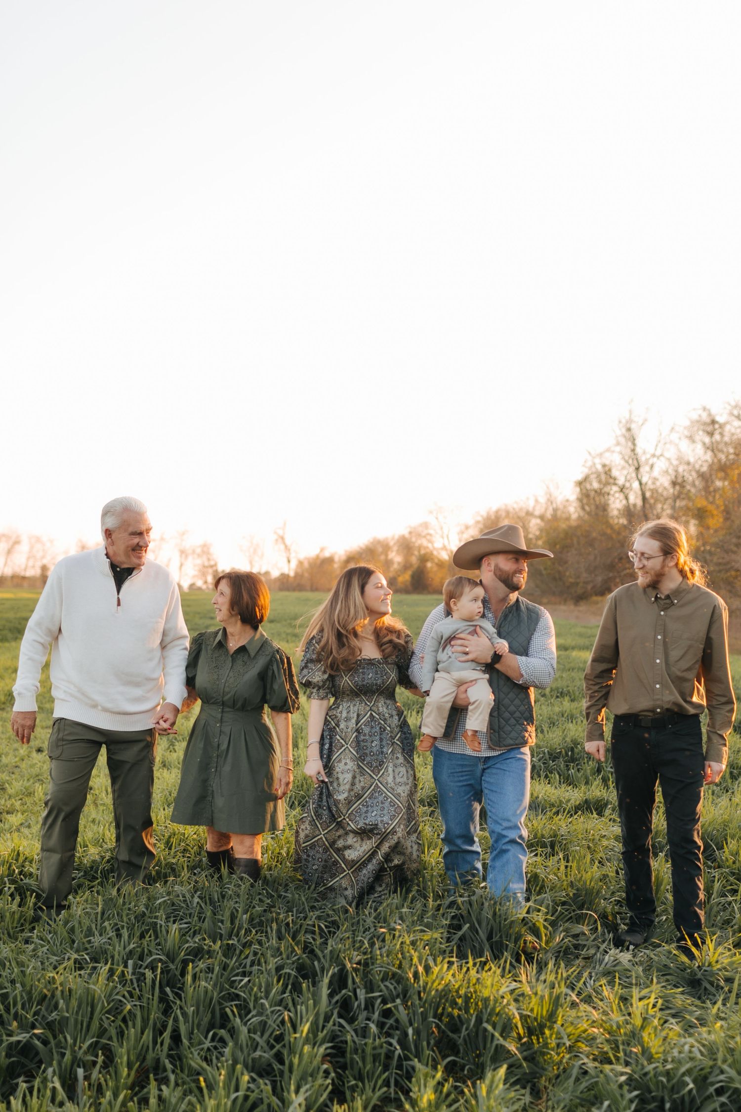 Extended family of six walking together through green field at golden hour in casual country attire.
