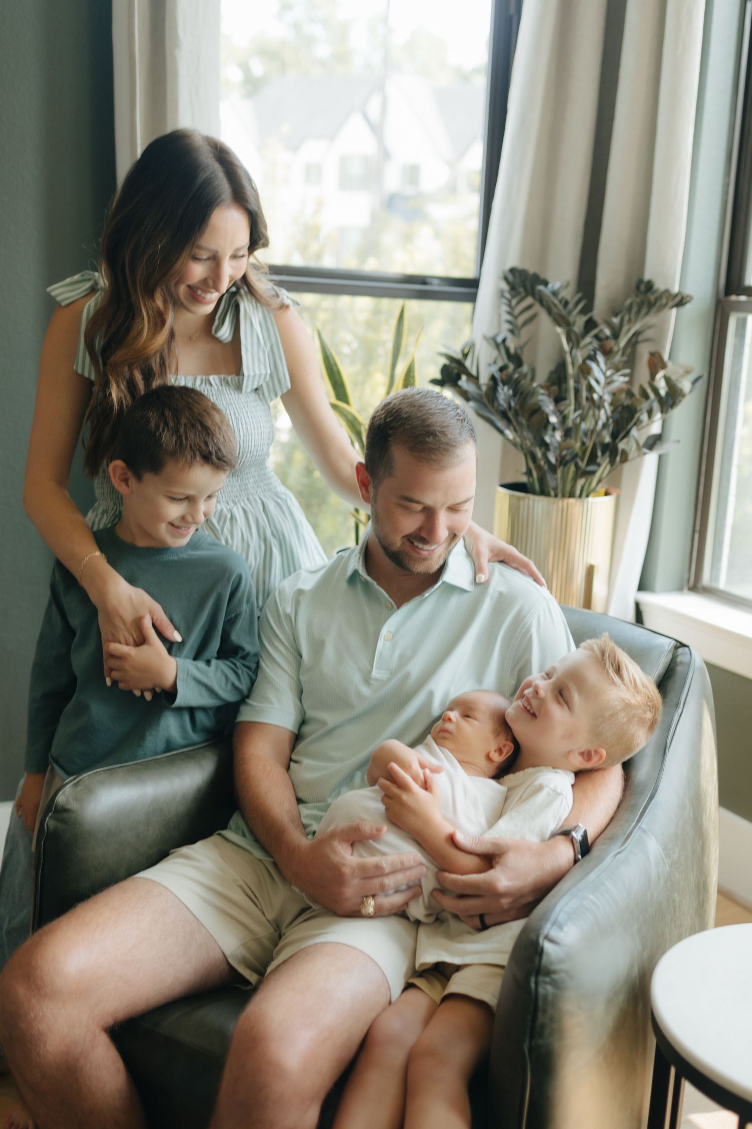 Smiling family of five with newborn baby gathered together in chair by sunlit window.