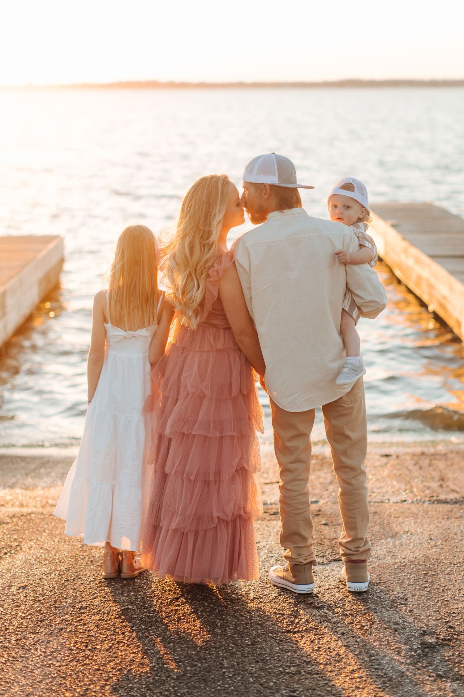 Family of four sharing a sunset kiss by the waterfront, with daughter in white dress and dad holding baby boy.