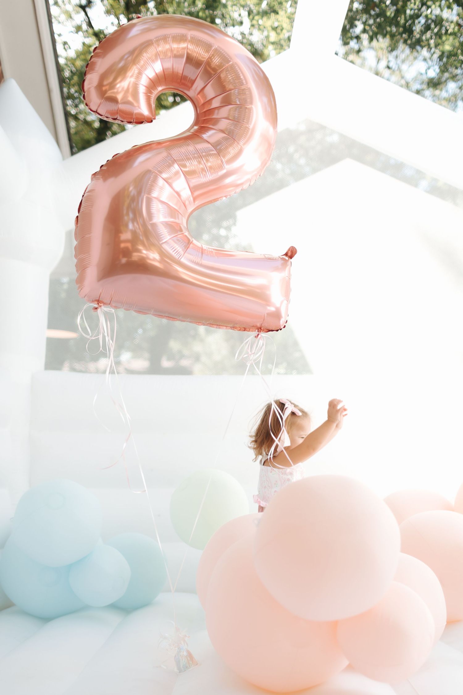 Toddler reaching for a rose gold number 2 balloon surrounded by pastel balloons at a 2nd birthday party.