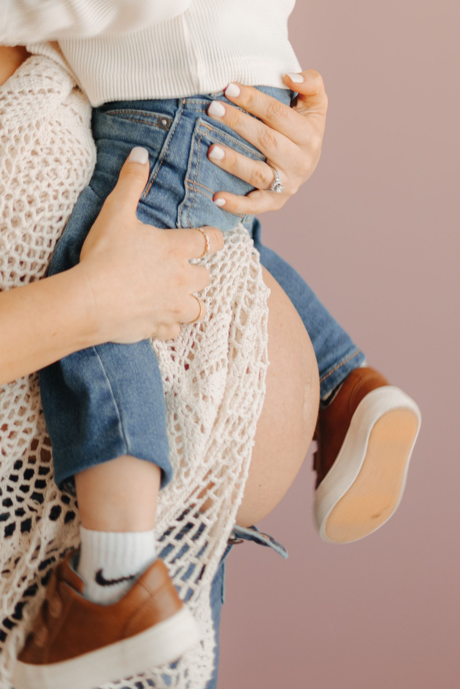 Pregnant woman in jeans and crochet cardigan holding a child wearing Nike socks and brown shoes against pink background.