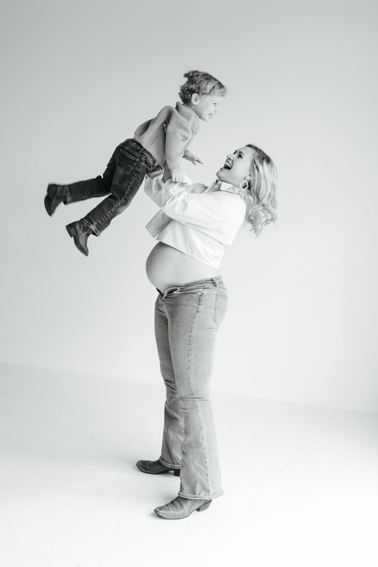 Black and white photo of a pregnant woman joyfully lifting her toddler son overhead in a bright studio.