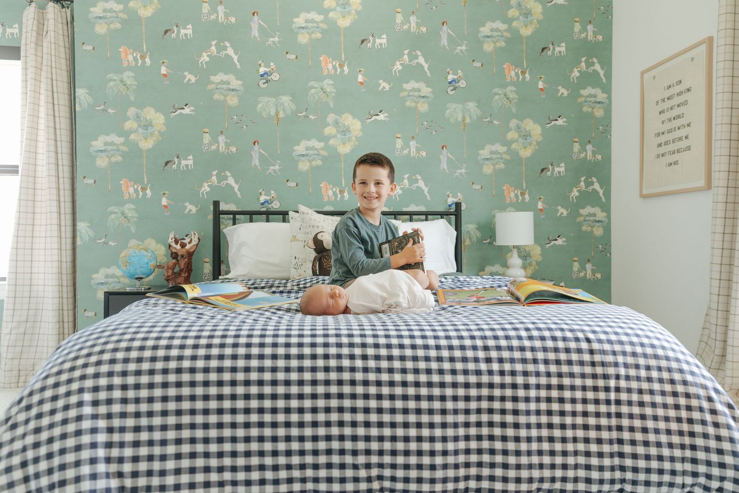 Young boy sits on gingham bedspread with newborn sibling in room with green toile wallpaper.