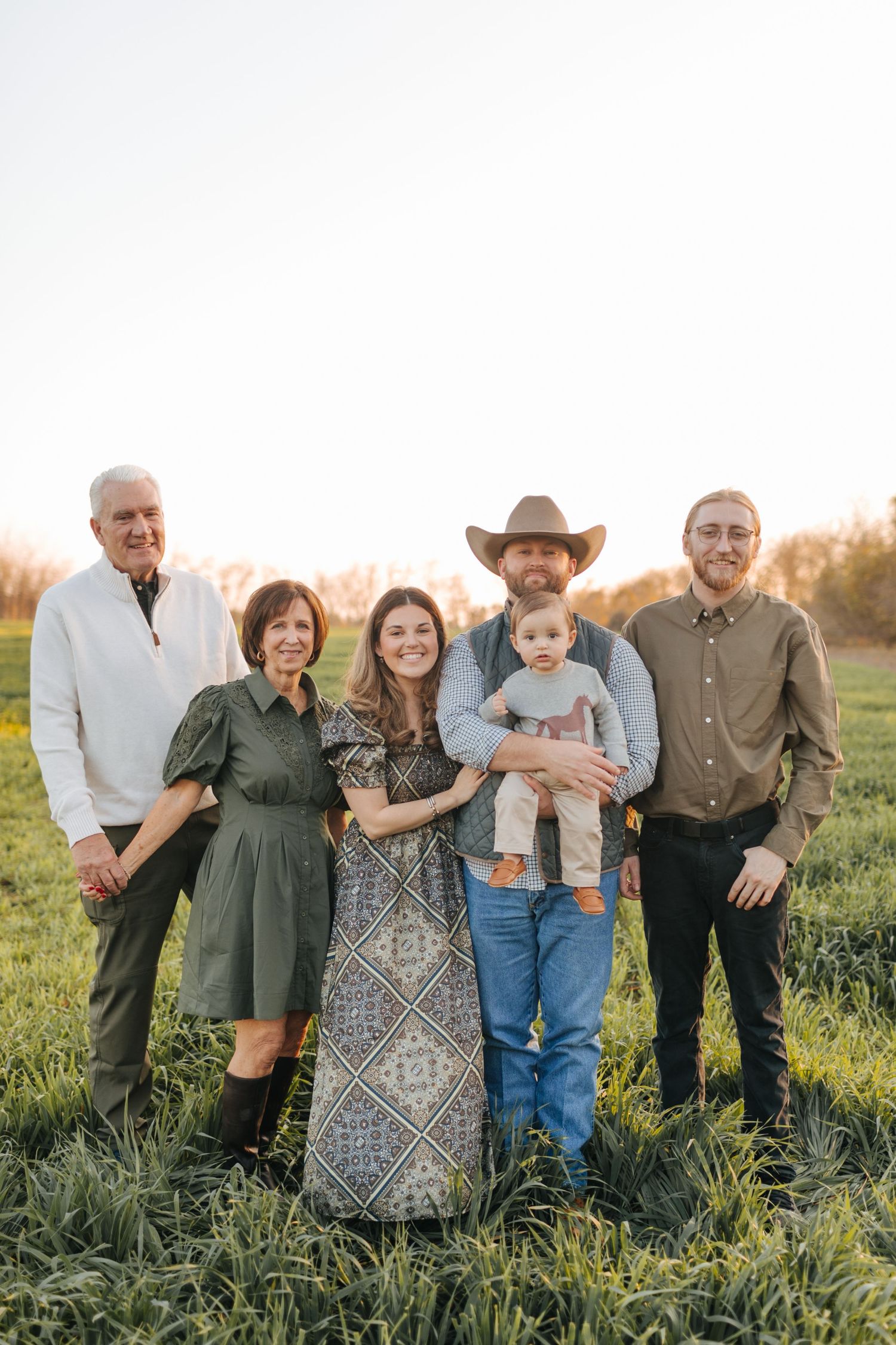 Multi-generational family of six poses together in lush green field during warm golden sunset light.