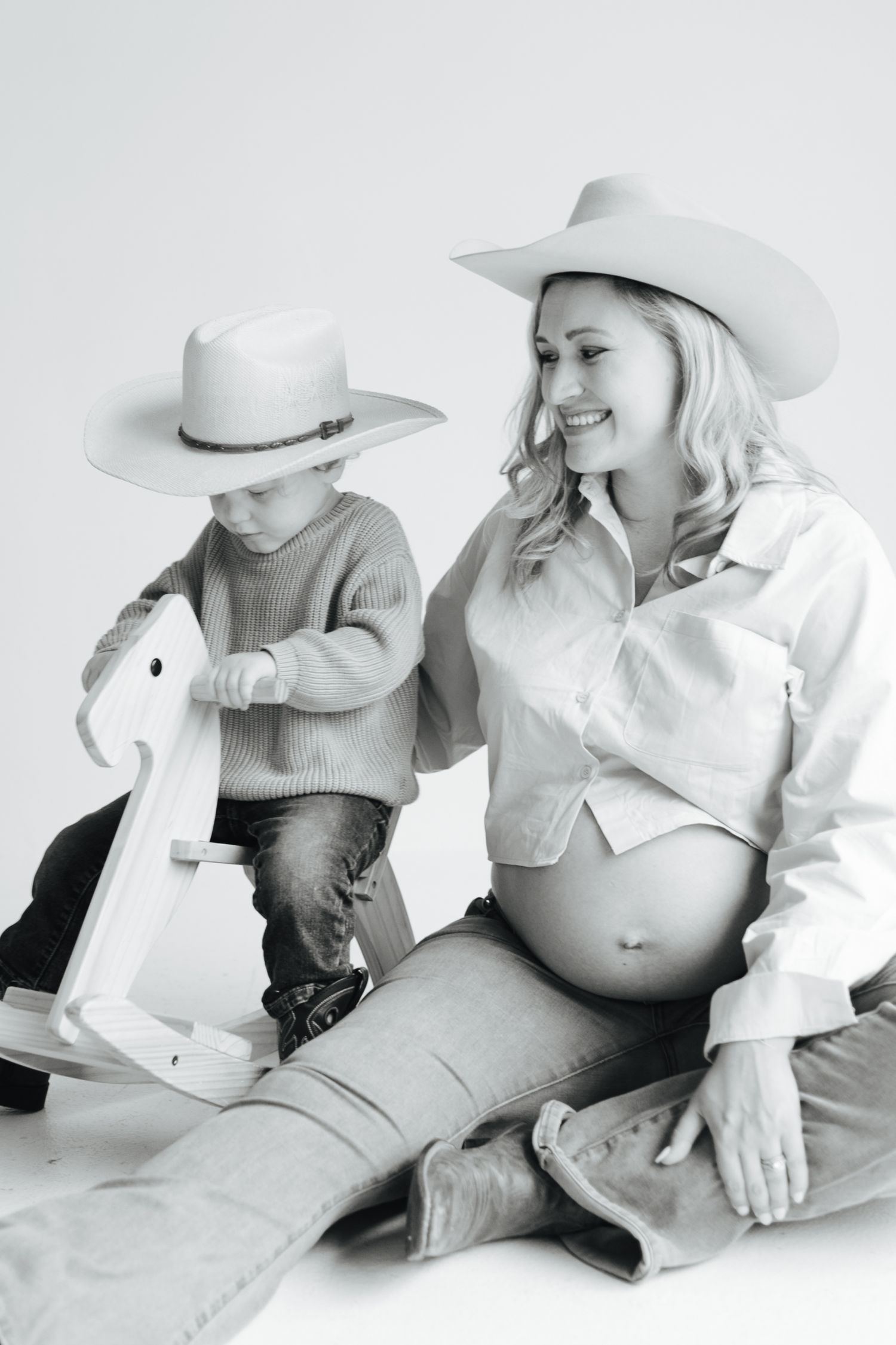 Black and white photo of pregnant woman and toddler in cowboy hats with a wooden rocking horse.