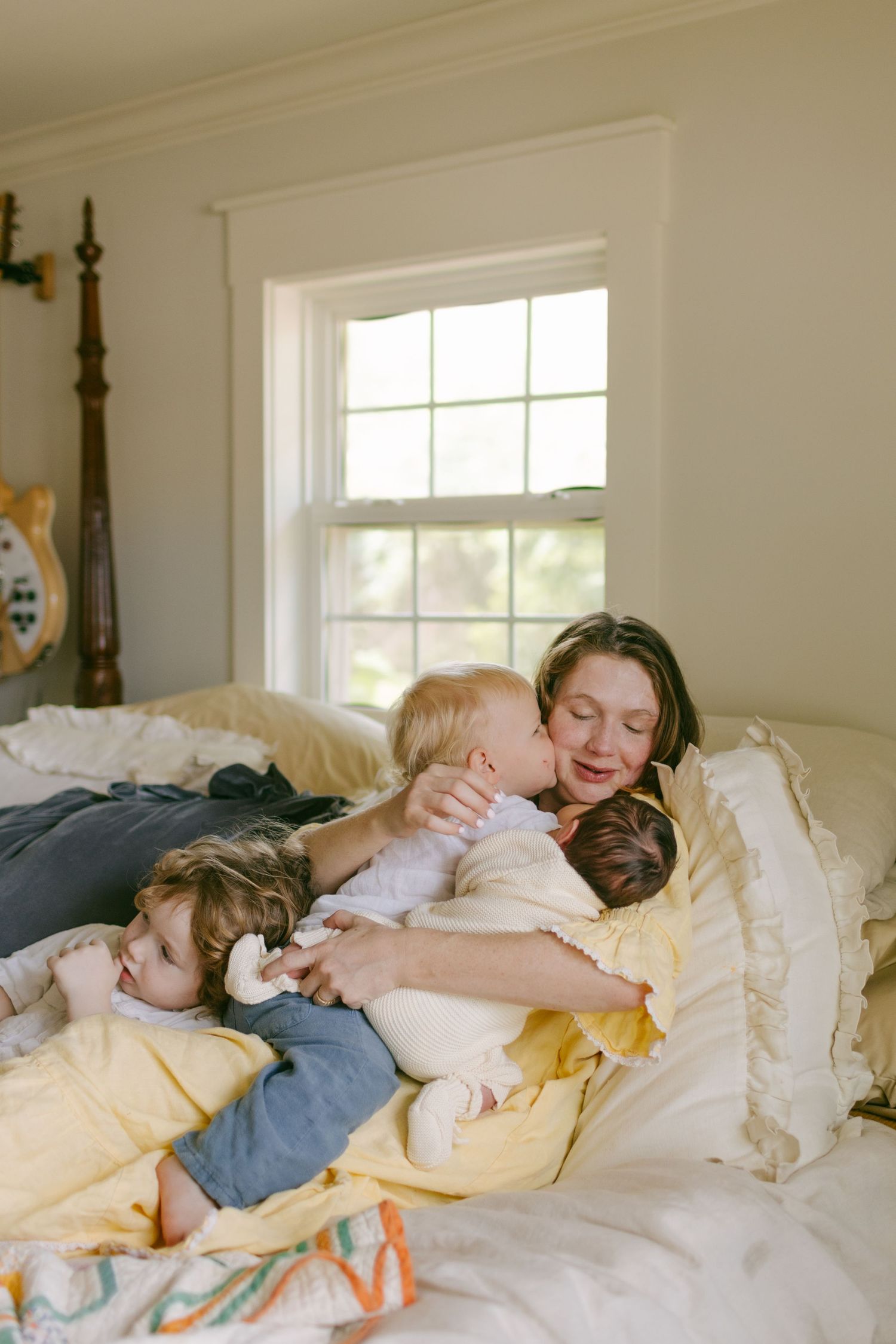 A mother cuddles her three young children on a bed with white bedding near a bright window.