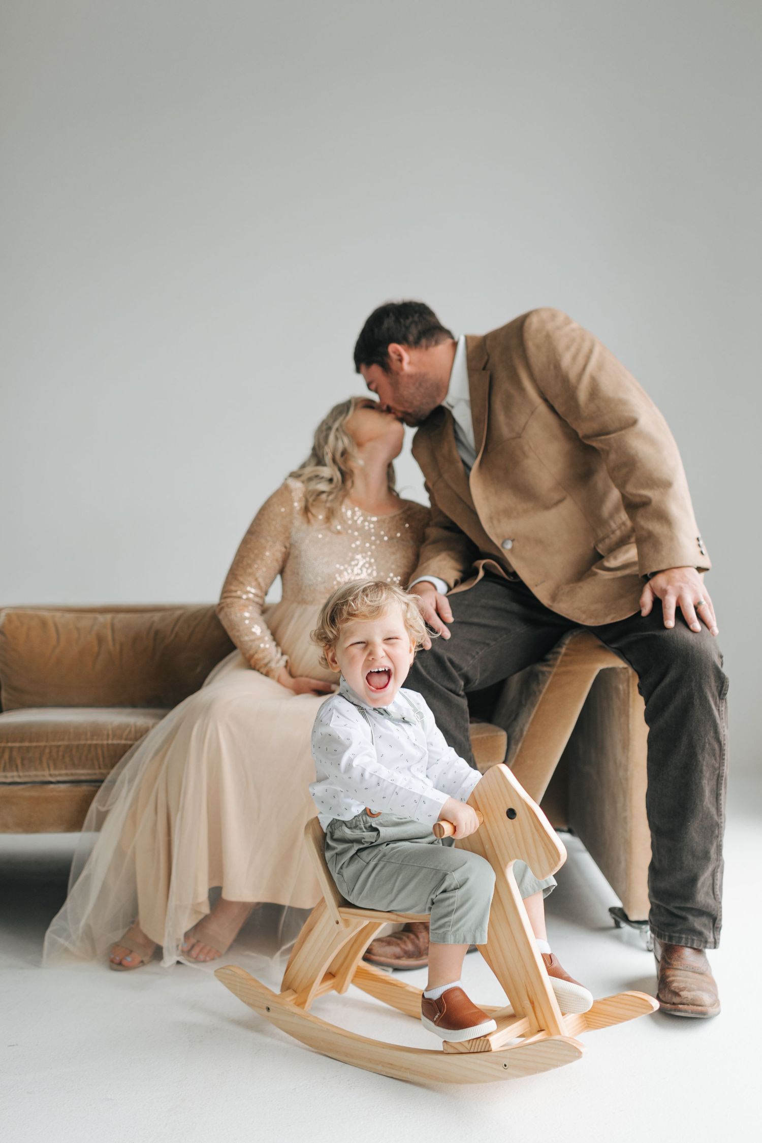 Parents kiss behind their giggling toddler son riding a wooden rocking horse in a bright studio.