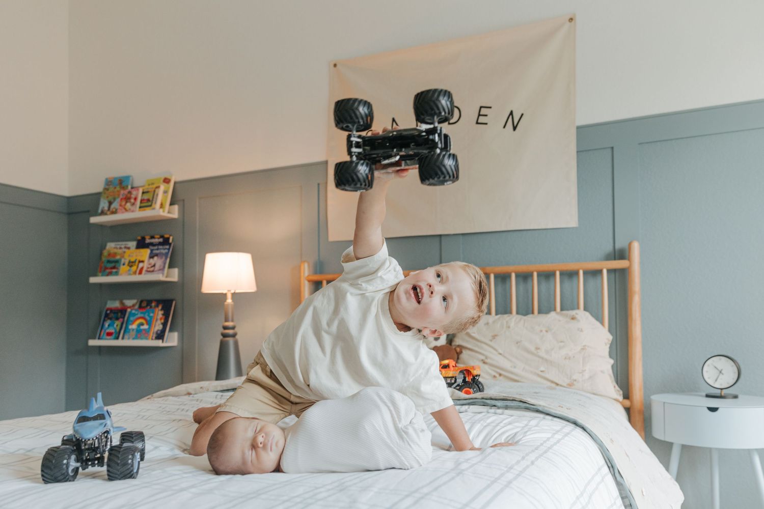 Toddler holds monster truck toy over smiling newborn baby on bed in blue and white nursery.