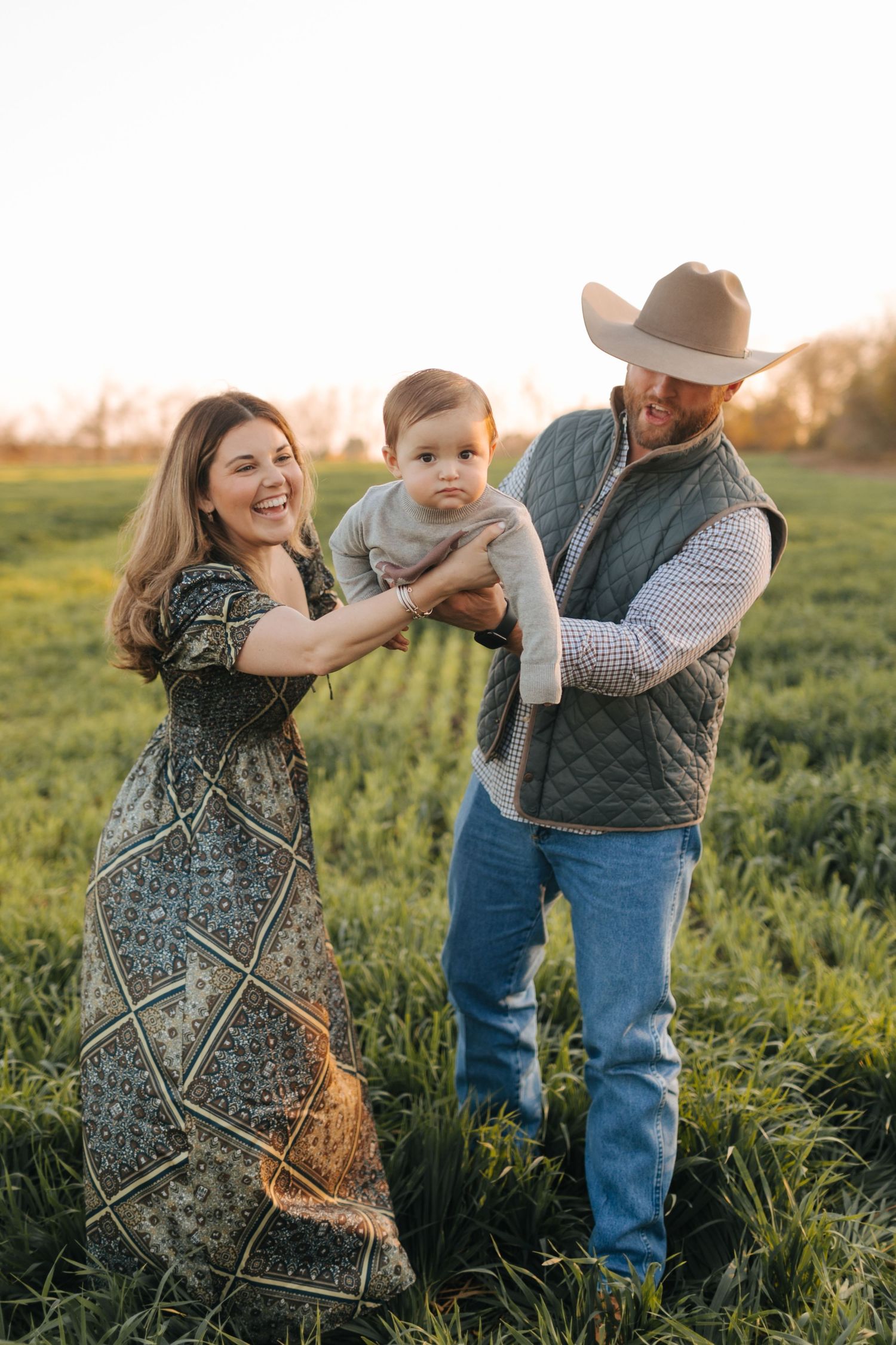 Young family with baby poses in green field at golden hour, mother in floral dress, father in cowboy hat and vest.