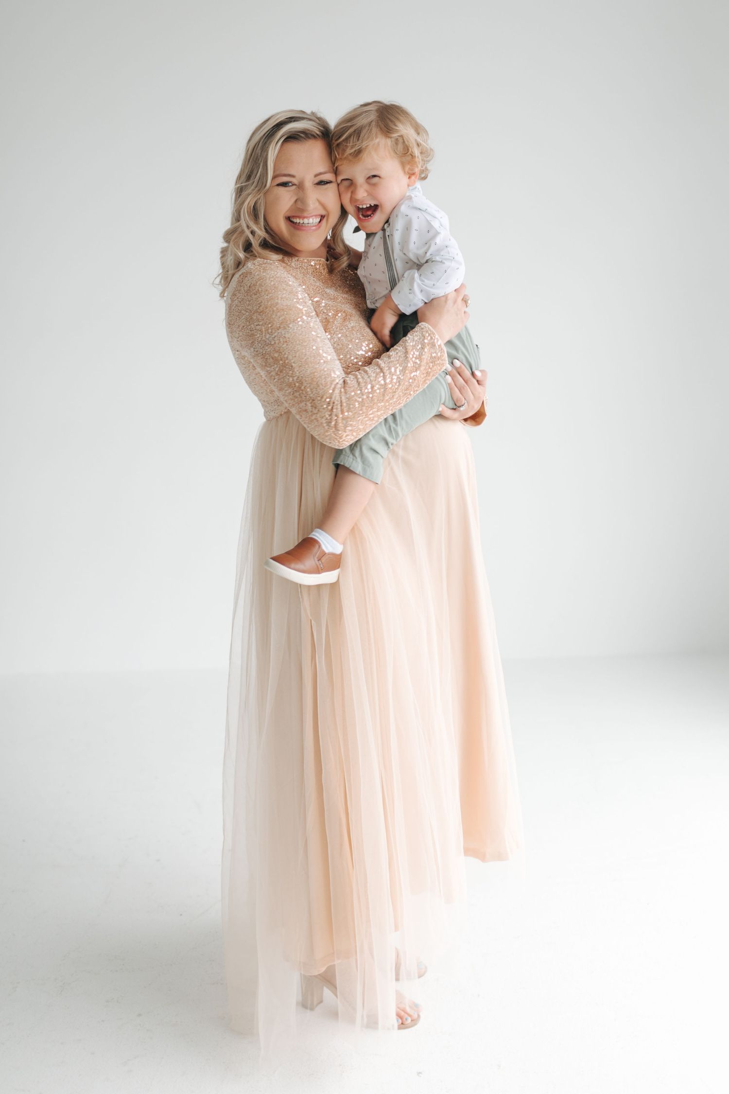 A smiling woman in a beige tulle gown holds a laughing toddler boy in a white studio setting.