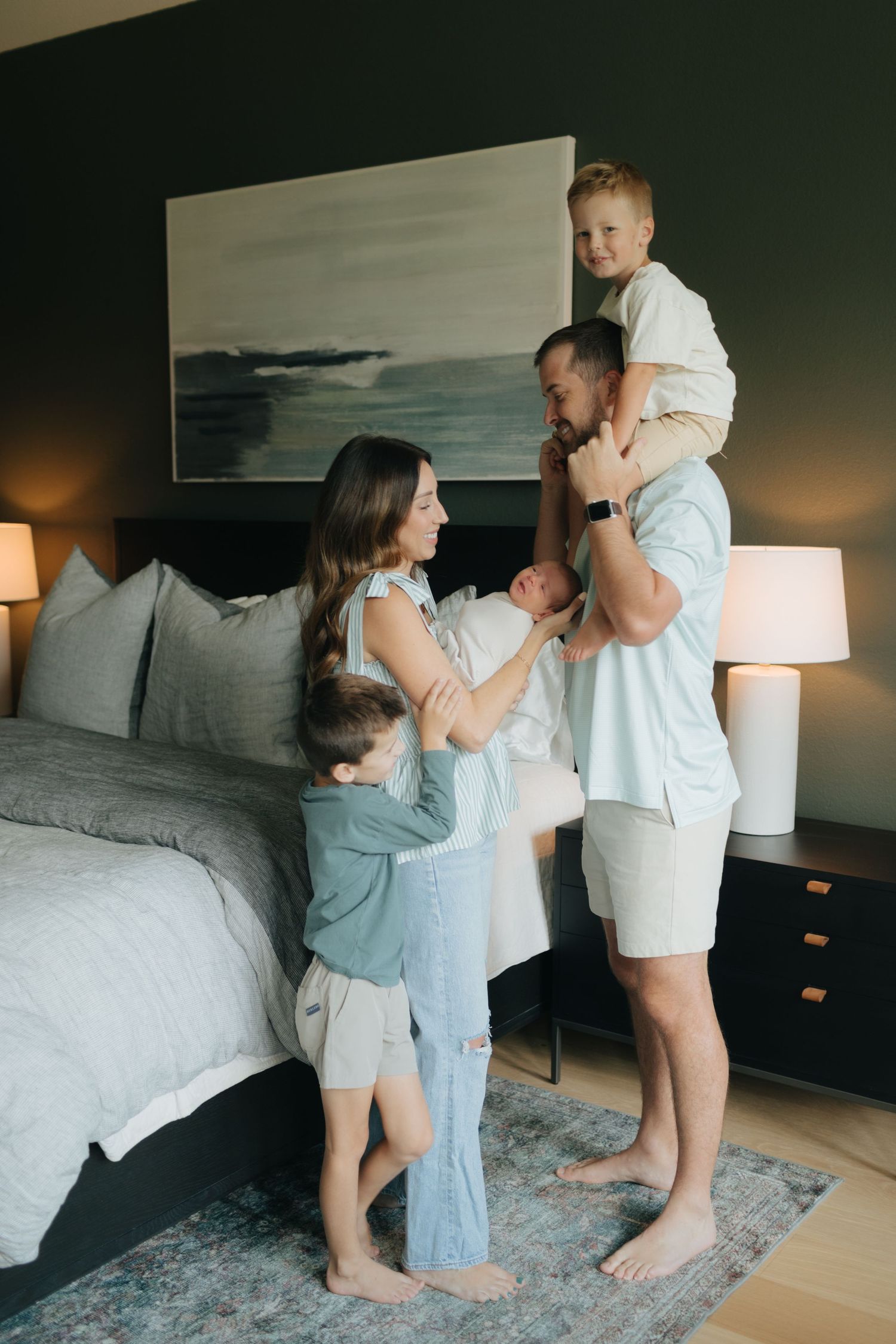 Family of five stands by bed, dad holding son on shoulders while mom holds newborn baby.