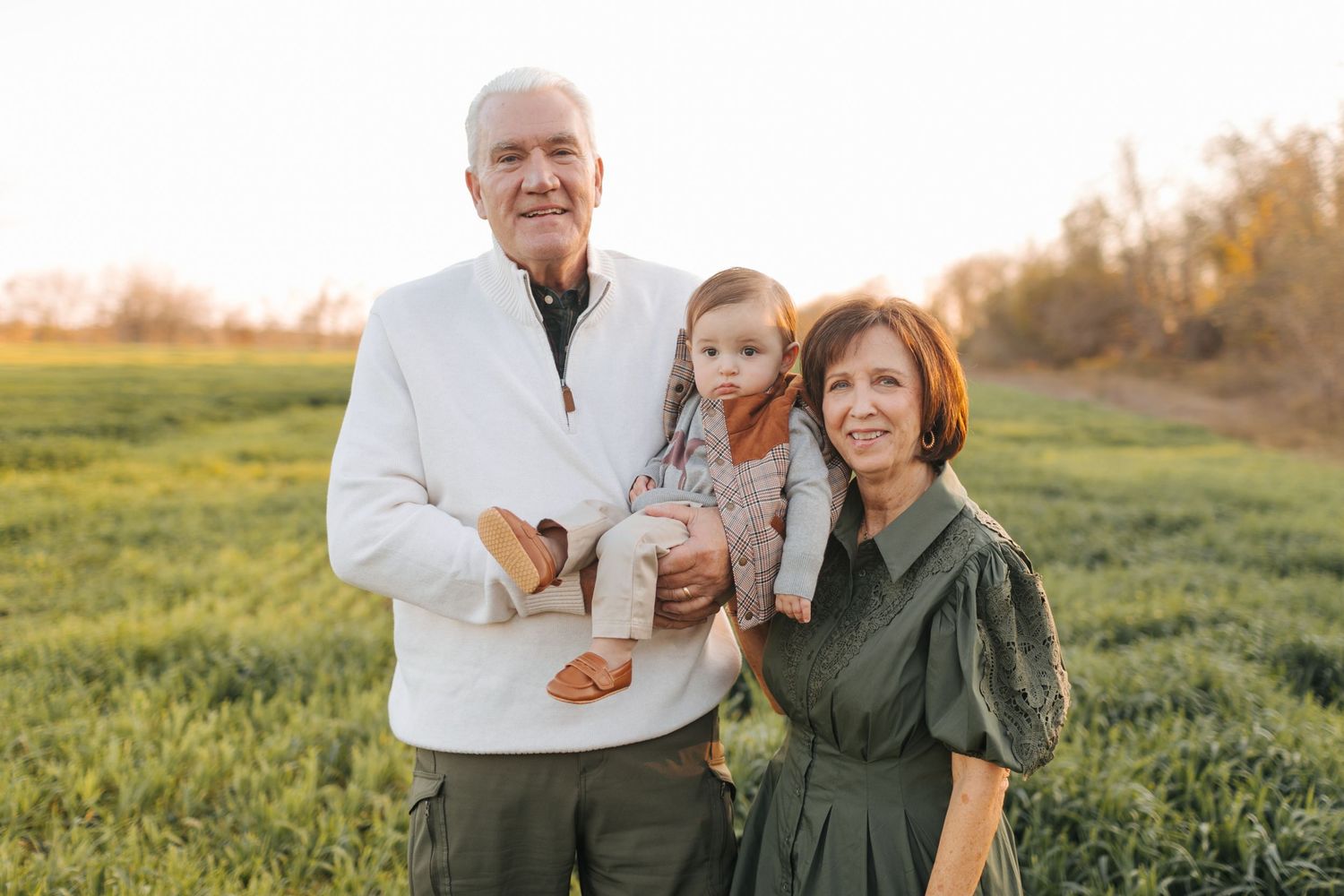 Happy grandparents holding a baby grandchild while standing in a green field at golden hour sunset.