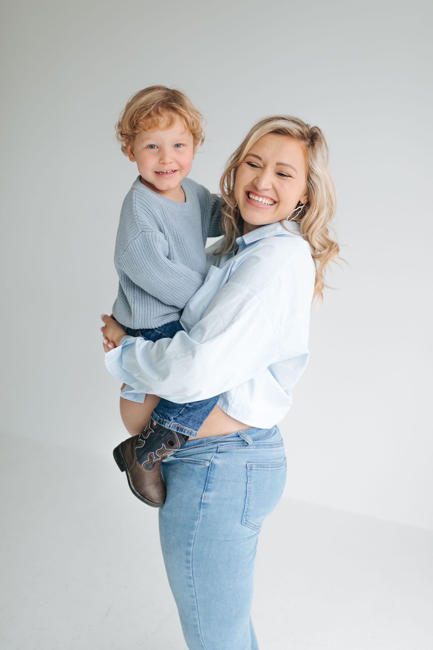 Smiling blonde mother holding her young redhead toddler son, both wearing blue outfits on white background.