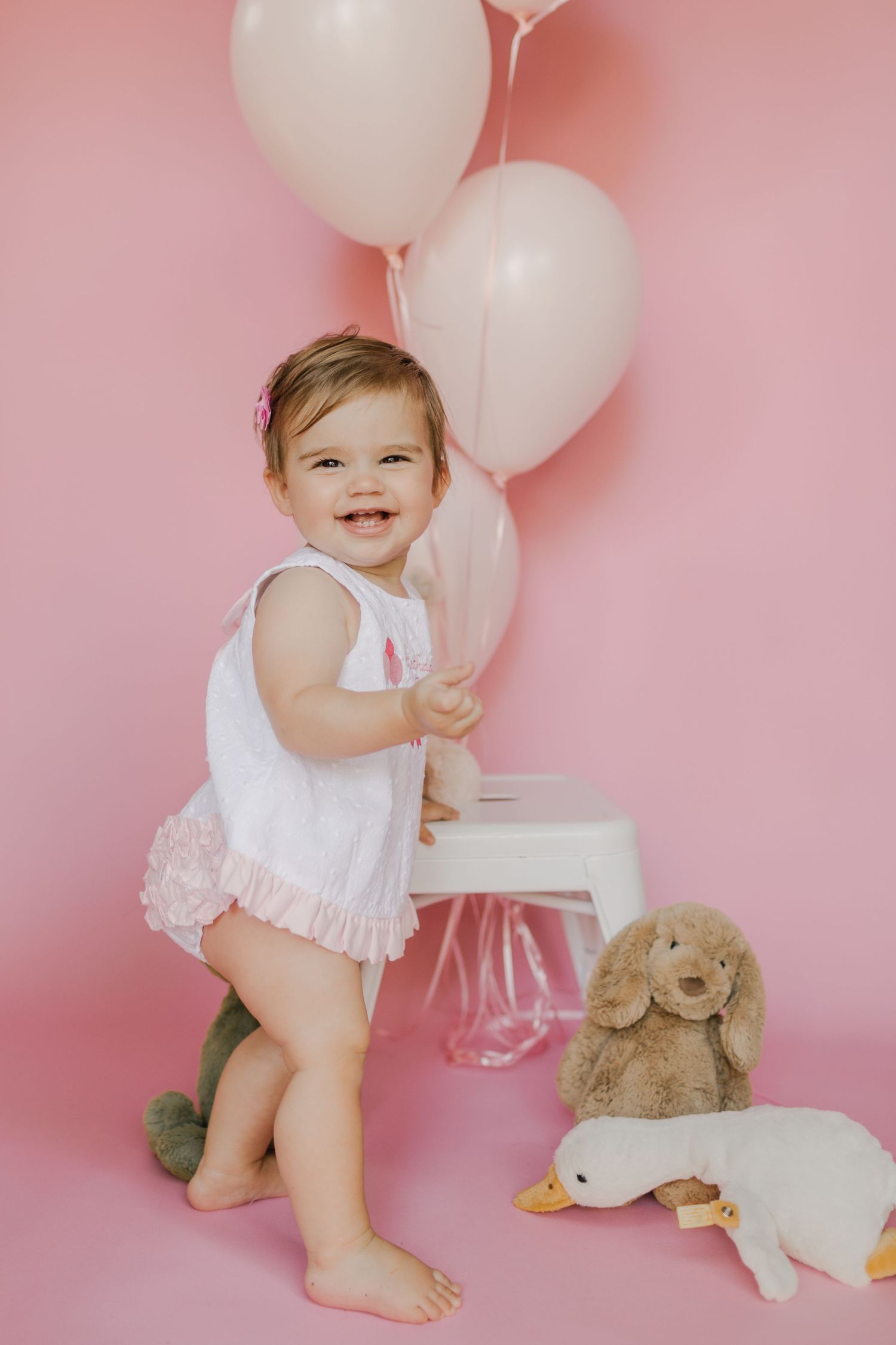 Smiling baby girl in white romper with pink balloons and stuffed animal toys on a pink background.