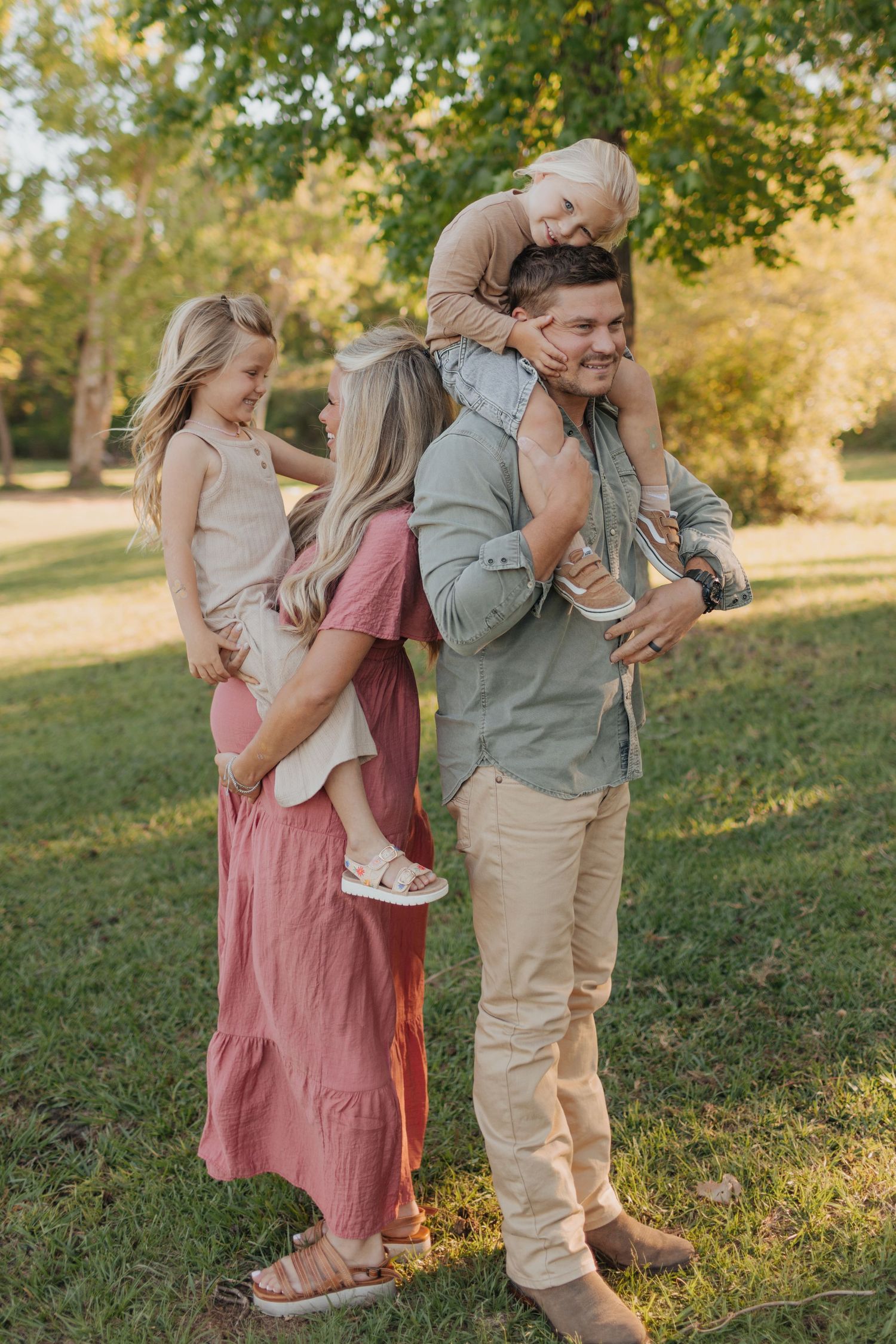 Joyful family of four standing back to back in park with kids riding on parents.