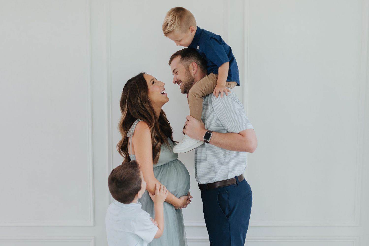 Pregnant woman in blue dress with husband holding toddler on shoulders and young boy beside them, family maternity photo.