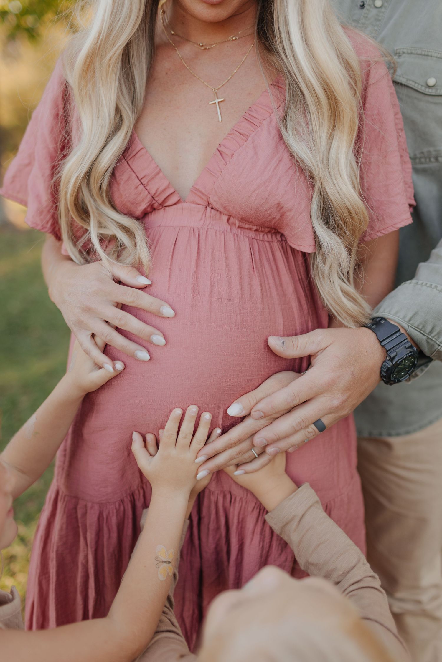Pregnant woman in pink dress with family hands touching baby bump during outdoor maternity photo session.