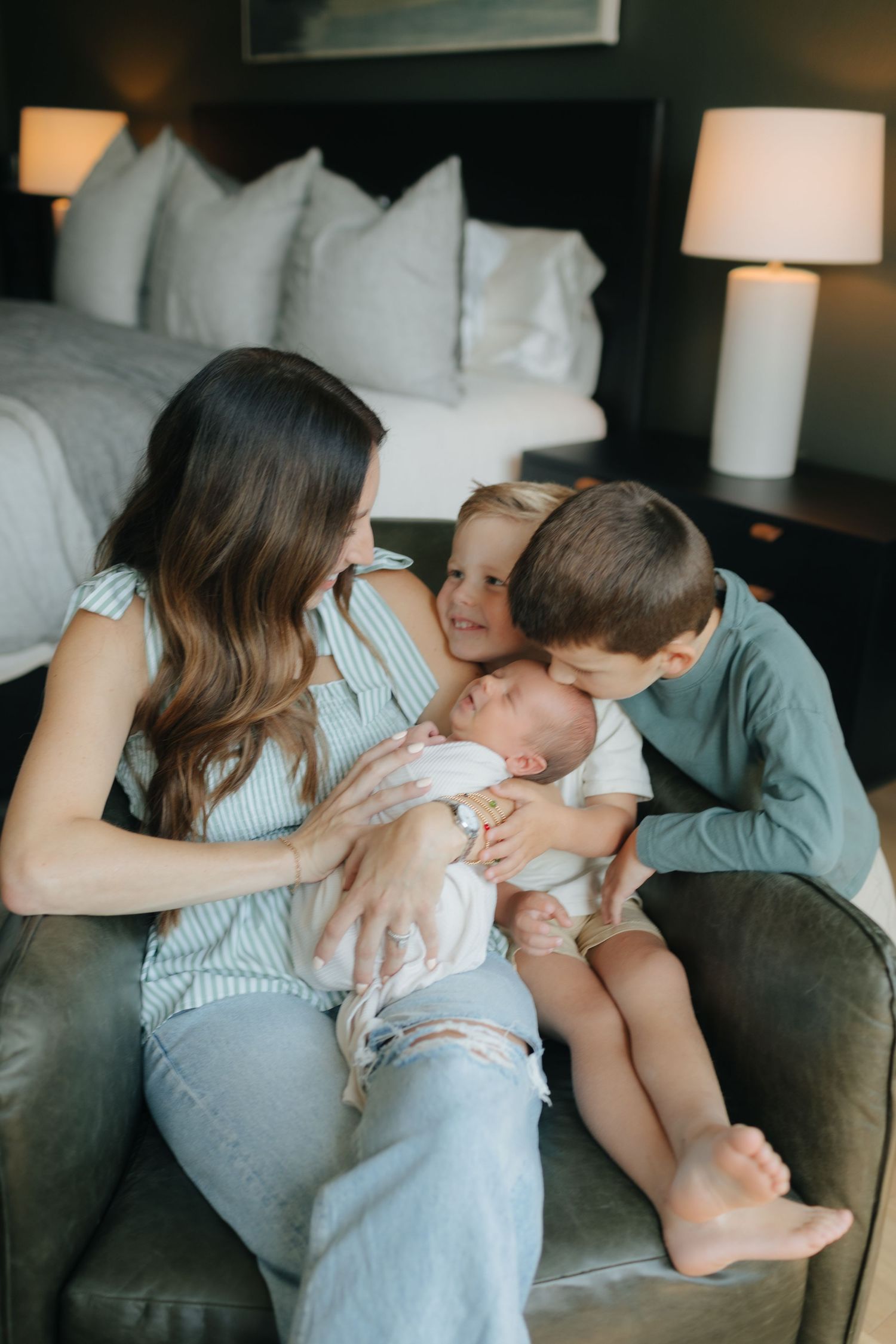 Mother sits in armchair holding newborn while two young siblings lean in to kiss the baby.