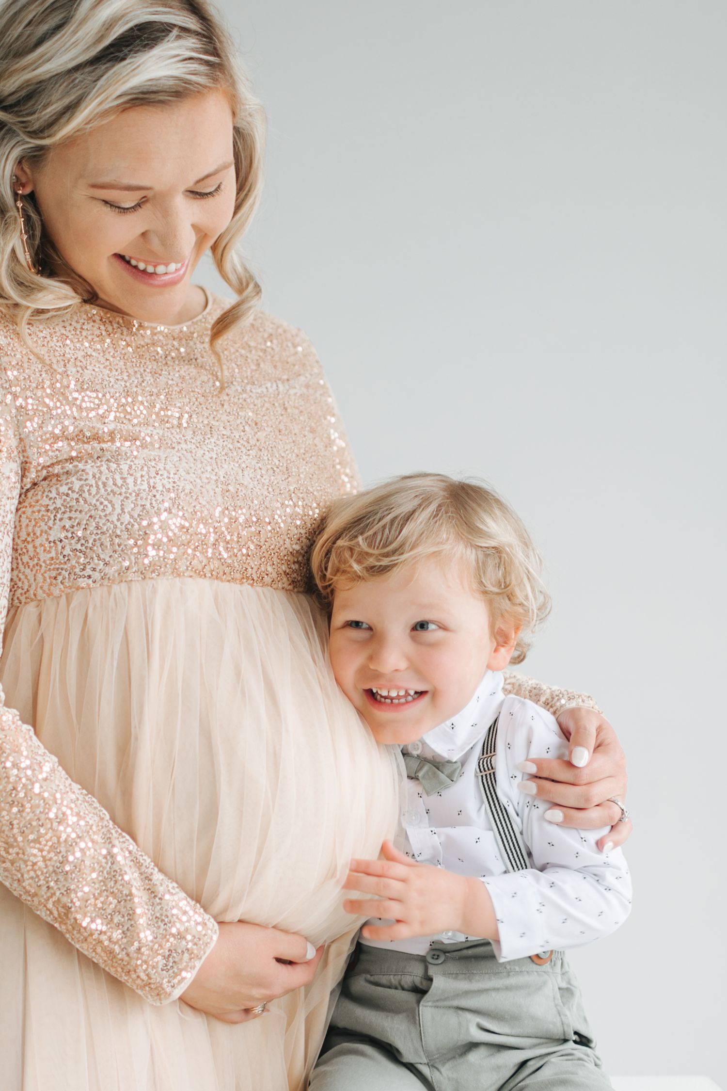 Smiling pregnant woman in sequin gown with young son hugging her baby bump.