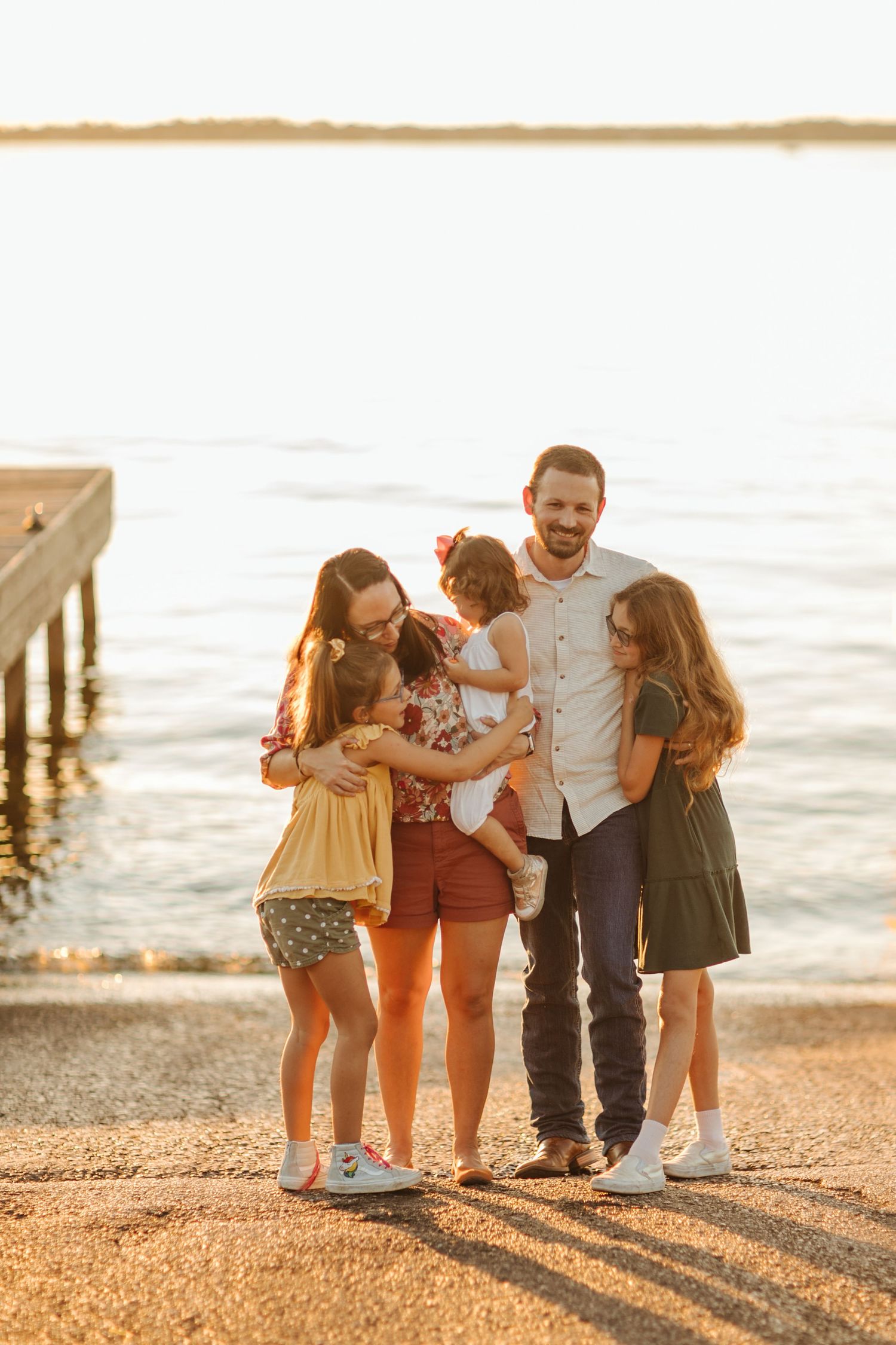 Family of five sharing a warm embrace on a sunlit lakeside dock during golden hour sunset.