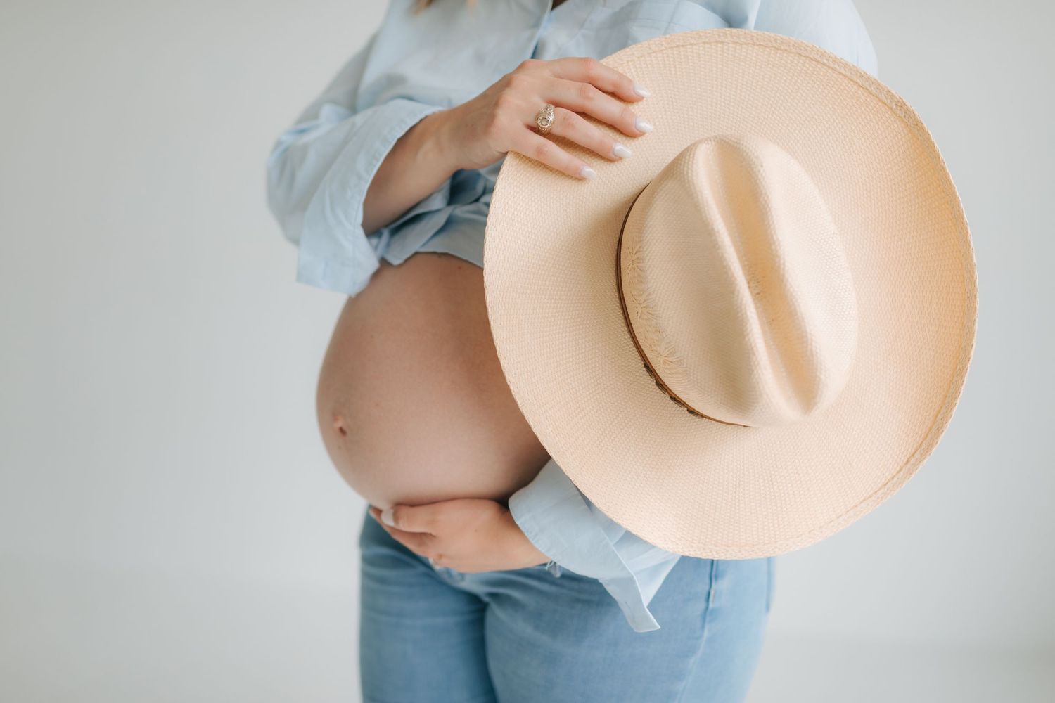 Close-up of pregnant woman holding a tan cowboy hat over her bare baby bump in a blue shirt.