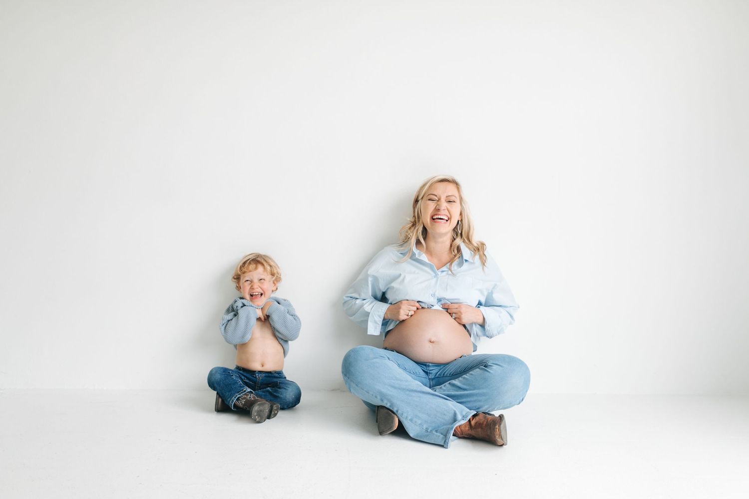 A laughing pregnant woman and her amused toddler son sit together in matching denim outfits in a white studio.