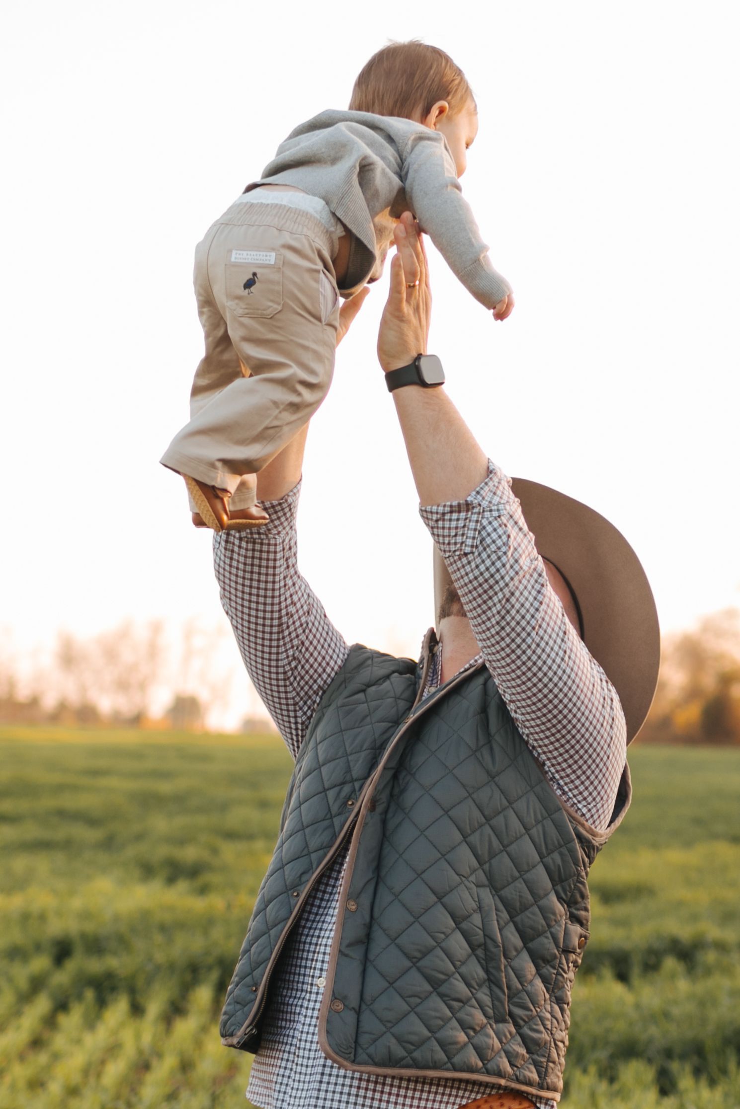 Father in cowboy hat and quilted vest lifts baby overhead in sunny green field at golden hour.