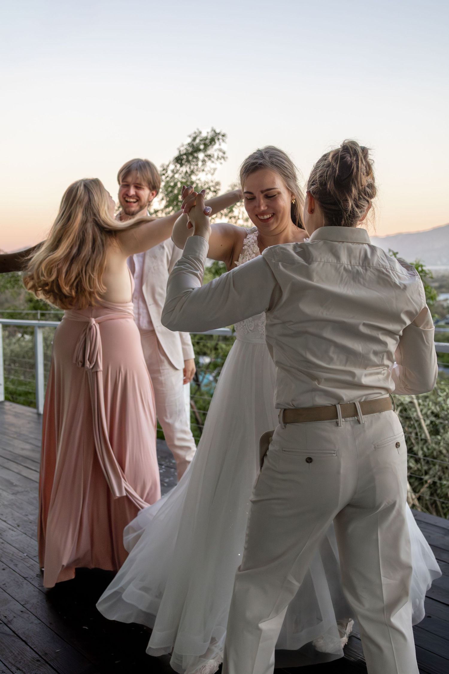 Bride in white gown dances joyfully with guests at outdoor wedding reception during golden hour sunset.