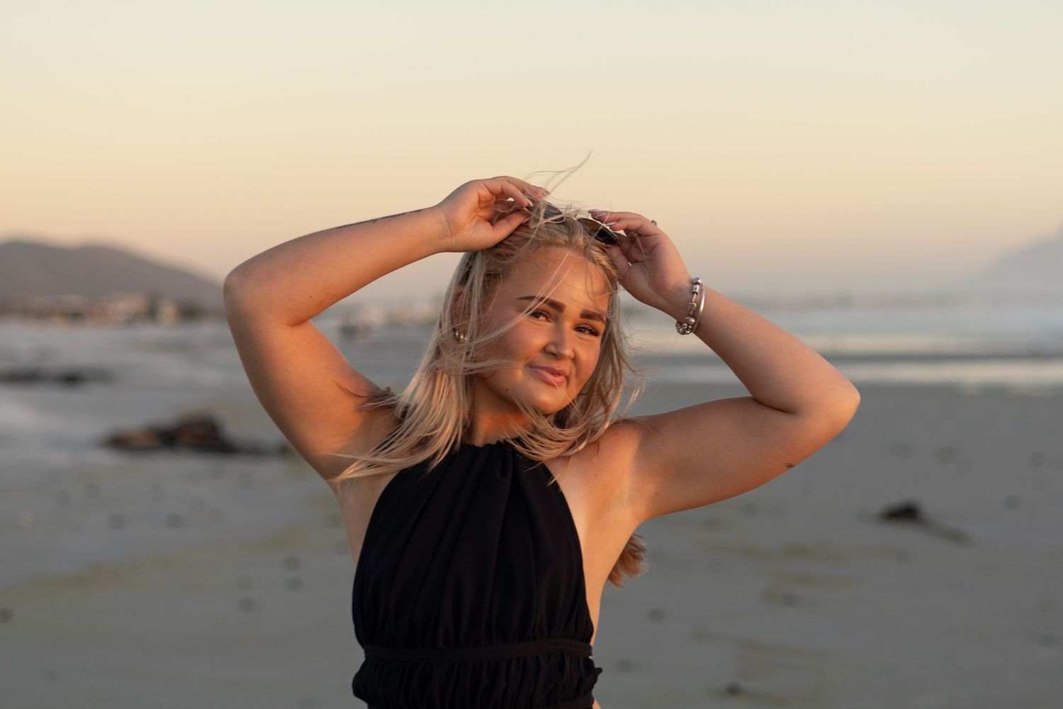 Smiling blonde woman in black halter top posing on beach during warm sunset.