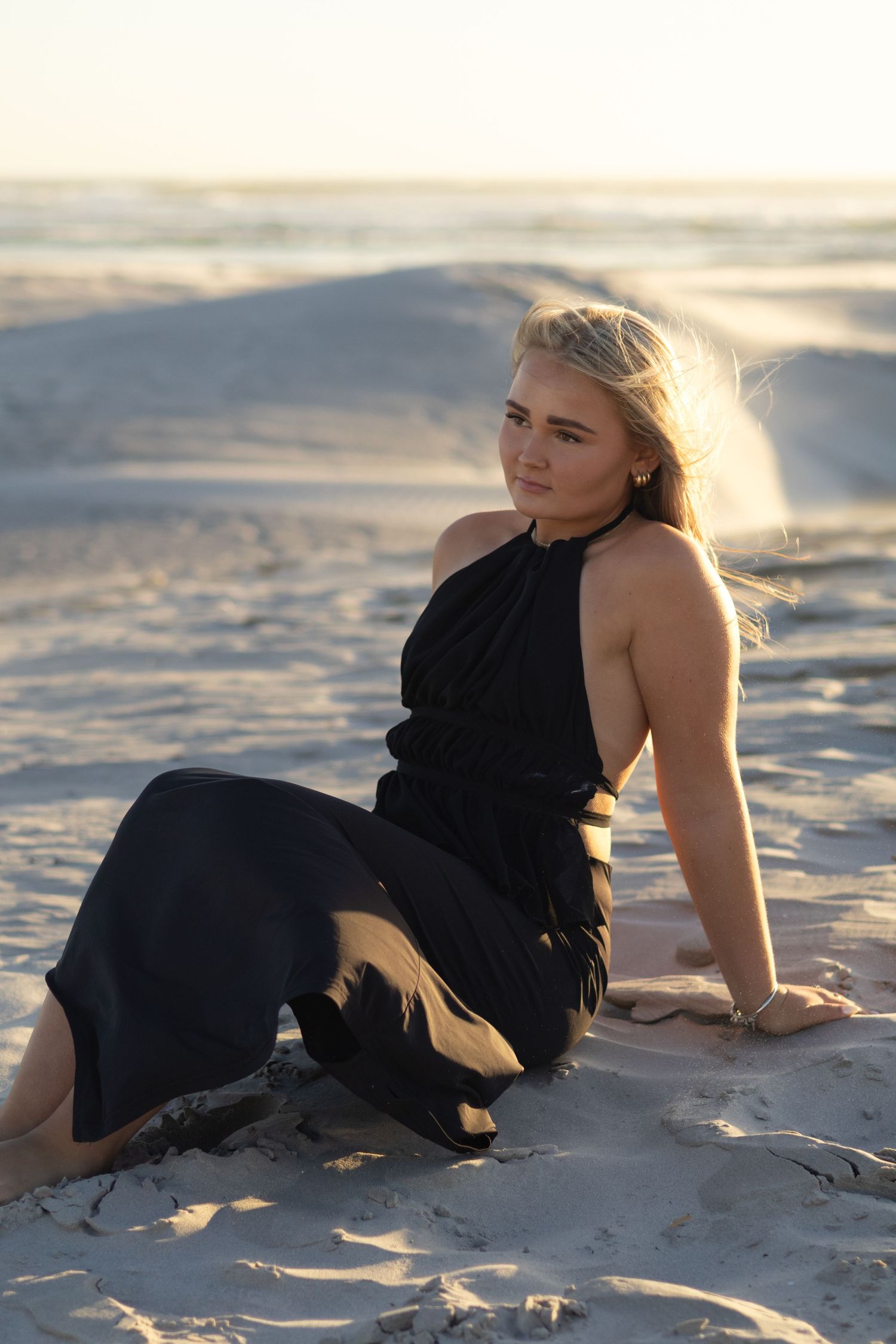 Young blonde woman in black halter dress sitting gracefully on sandy beach at golden hour sunset.