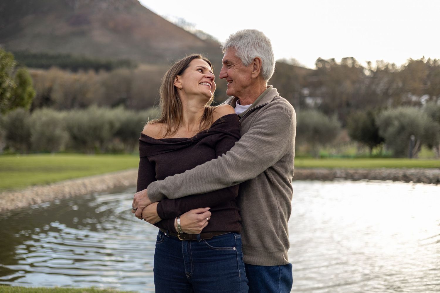 Happy mature couple embracing and smiling by a scenic pond with mountains and trees in the background.